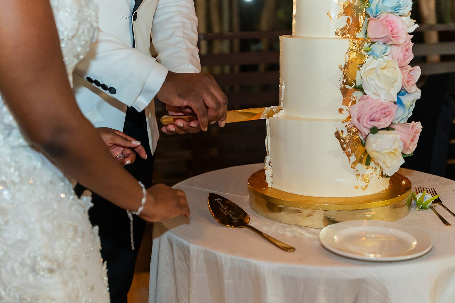Bride and groom cutting a elegant white wedding cake adorned with pastel flowers and gold accents.