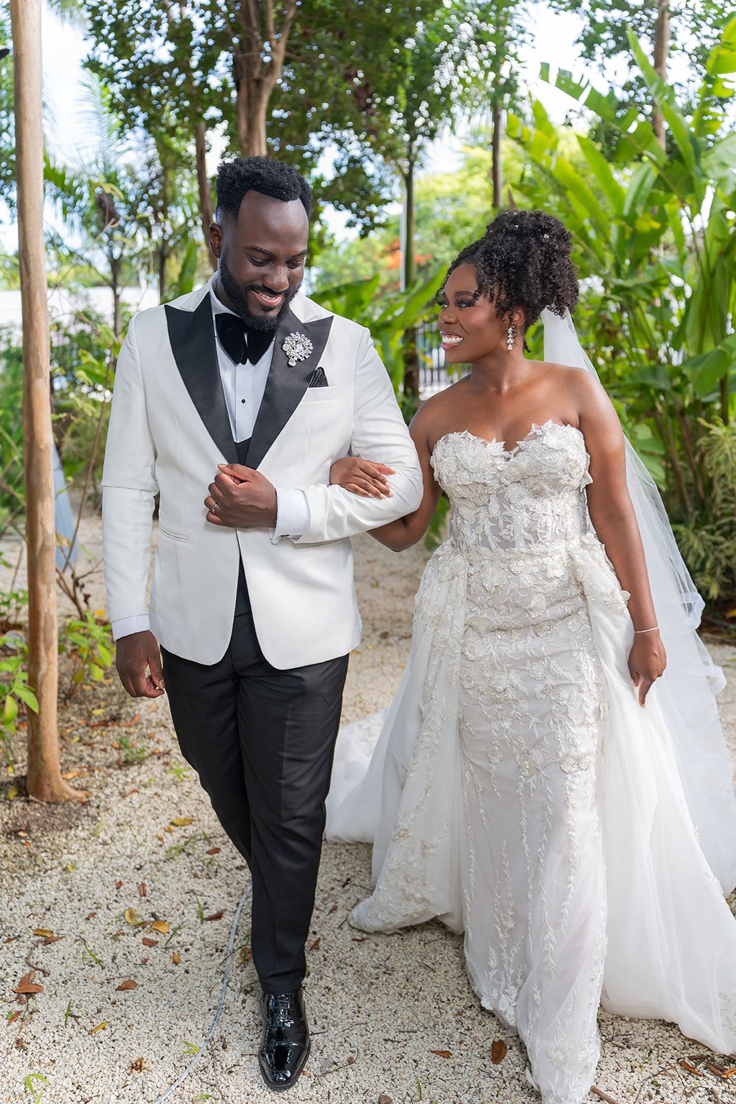 A smiling bride and groom walk arm in arm outdoors, she in a lace gown, he in a white tuxedo jacket.