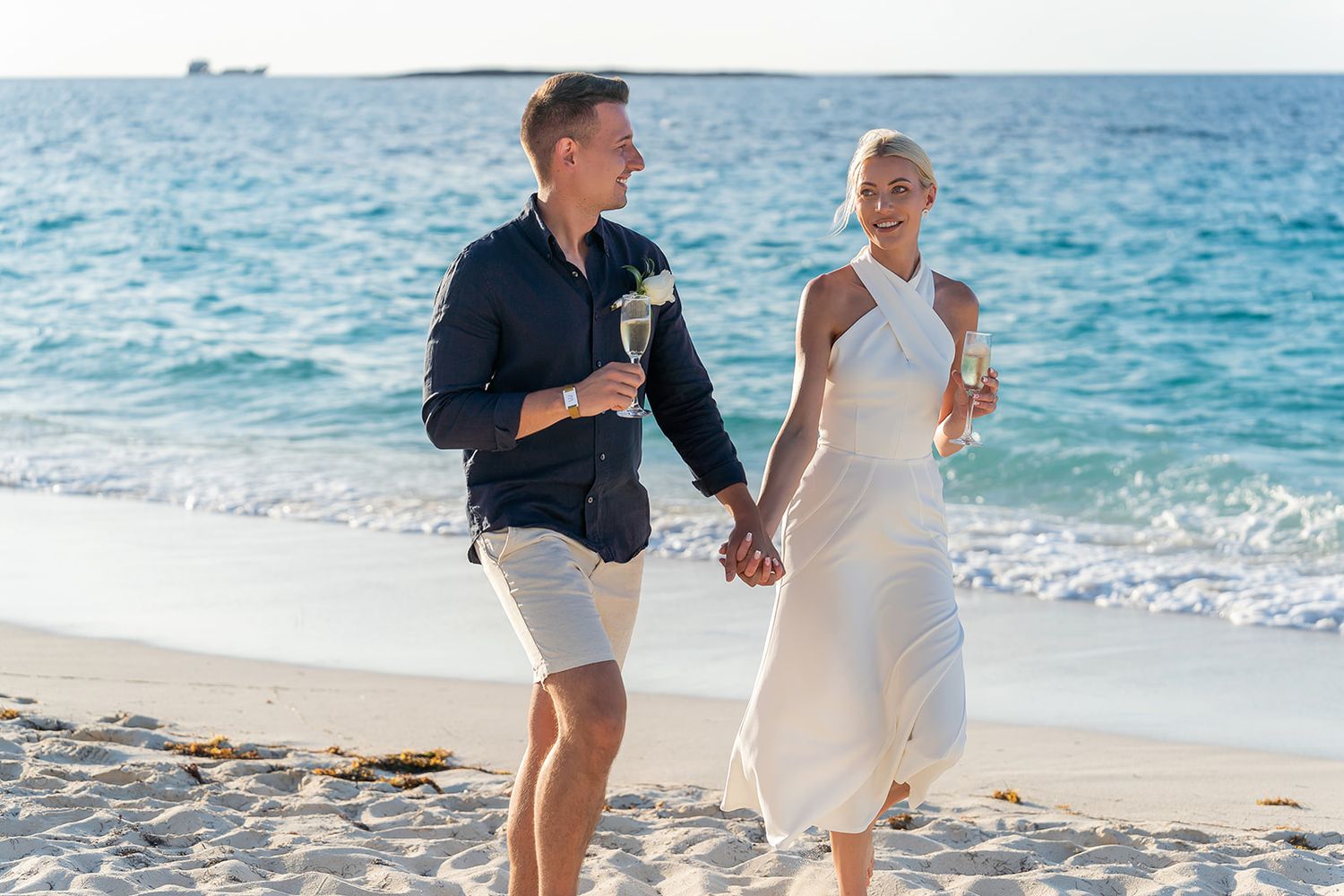 Couple holding champagne glasses and walking hand in hand on a sunny beach with turquoise ocean waves.