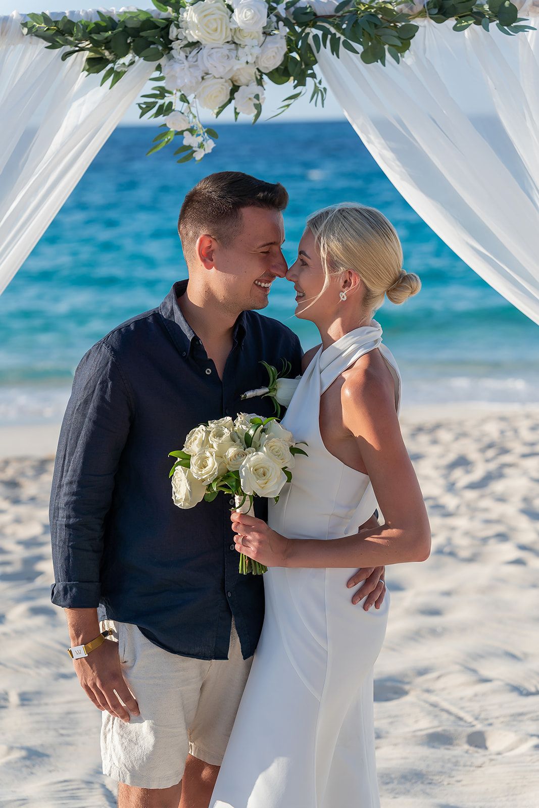 Couple sharing a romantic moment at a beach wedding ceremony under a white floral arch with ocean backdrop.
