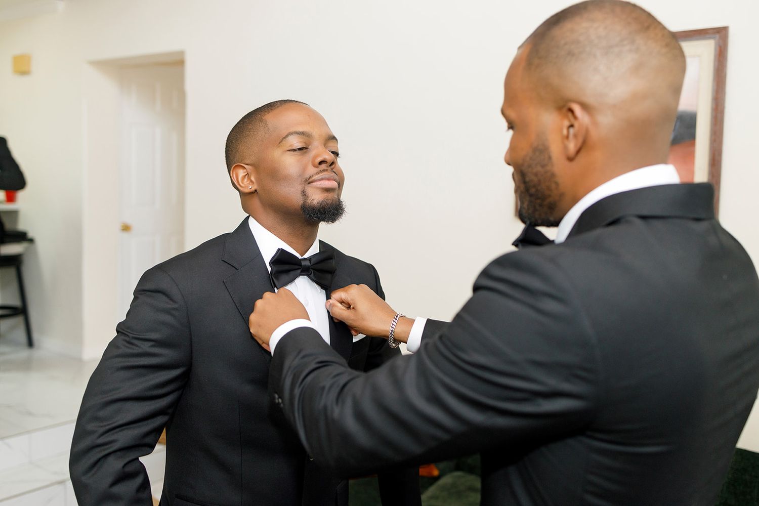 Two men in tuxedos, one adjusting the other's bow tie, preparing for a formal wedding event.
