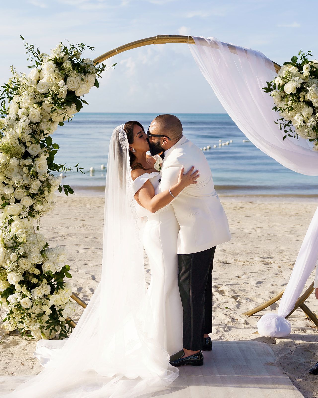Bride and groom share first kiss under floral arch on beach wedding ceremony.