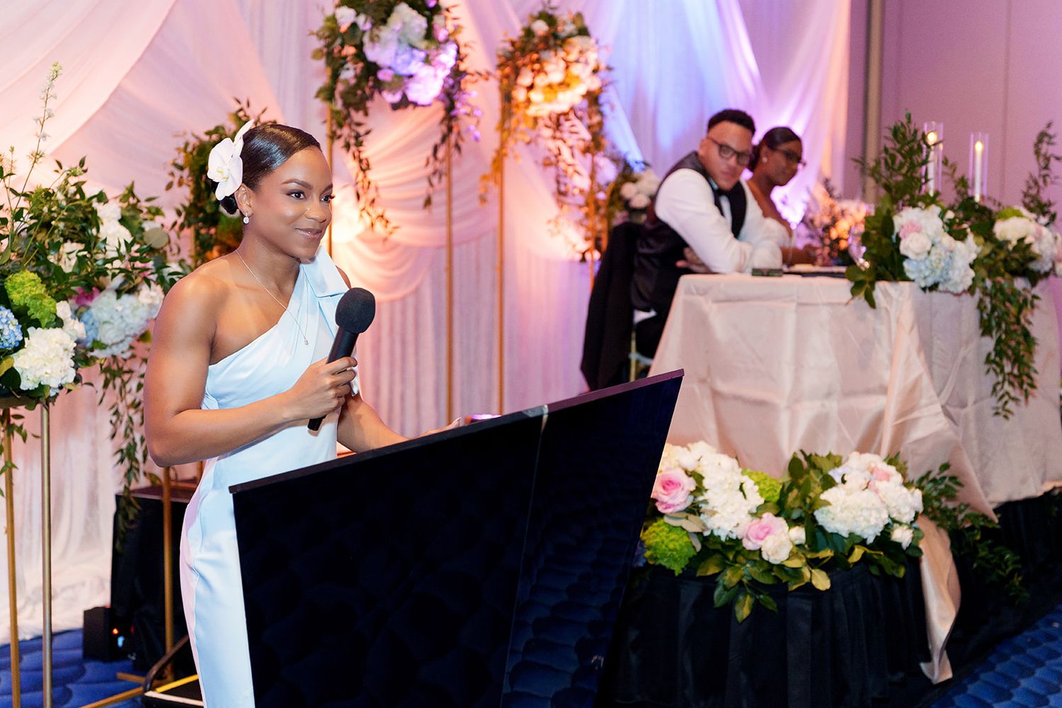 Woman in white dress speaks at podium with microphone at elegant floral wedding reception venue.