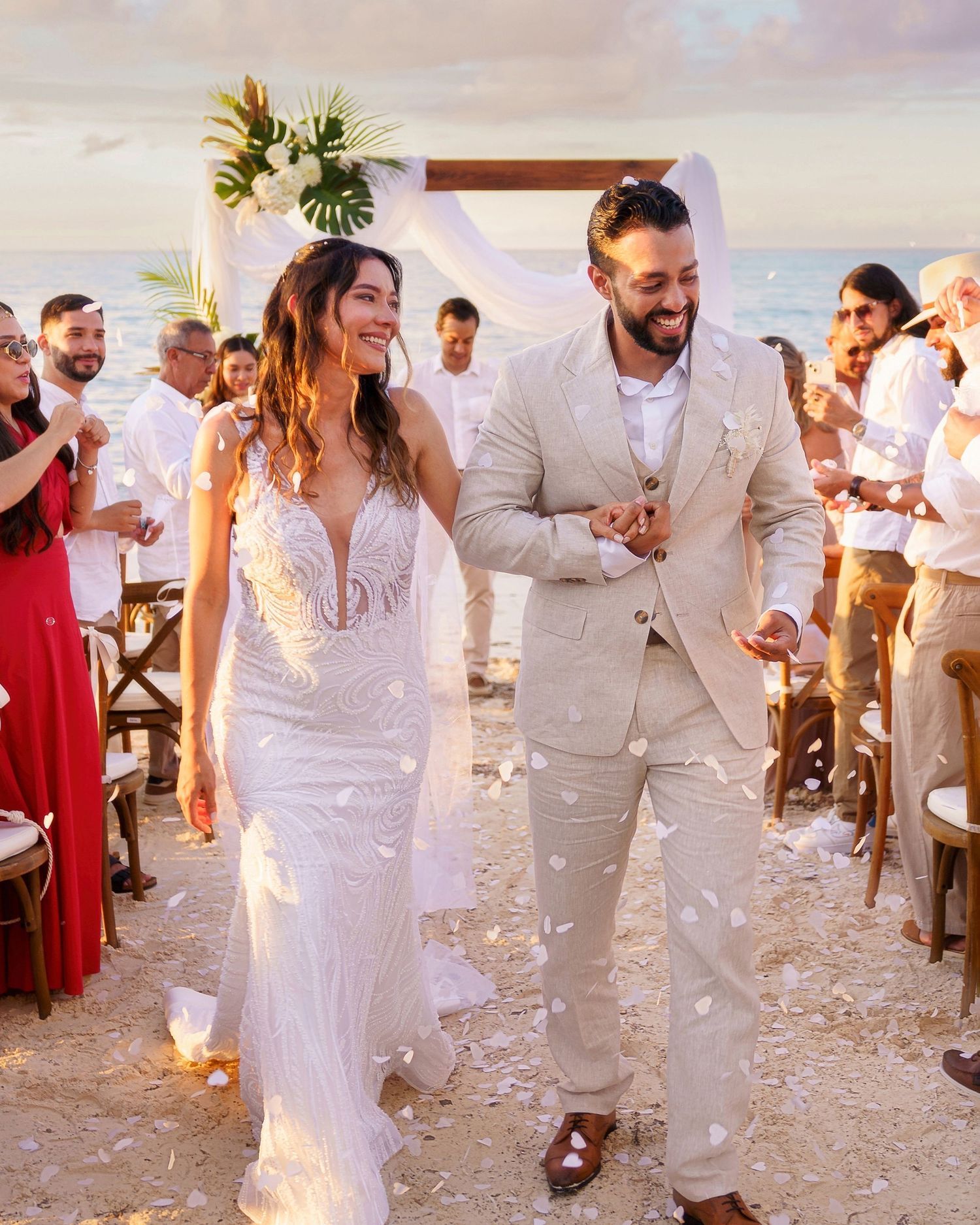 Newlywed couple walking down beach aisle in white dress and beige suit, with guests tossing white petals at sunset.