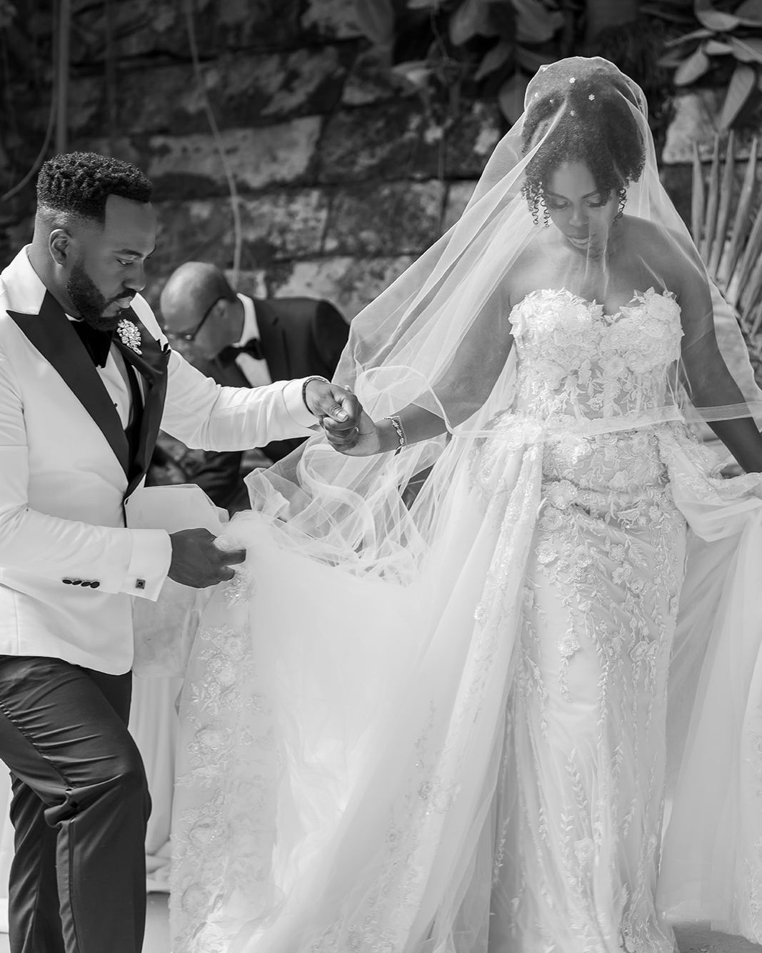 Black and white wedding photo of groom lifting bride's veil during outdoor ceremony, bride in lace gown.
