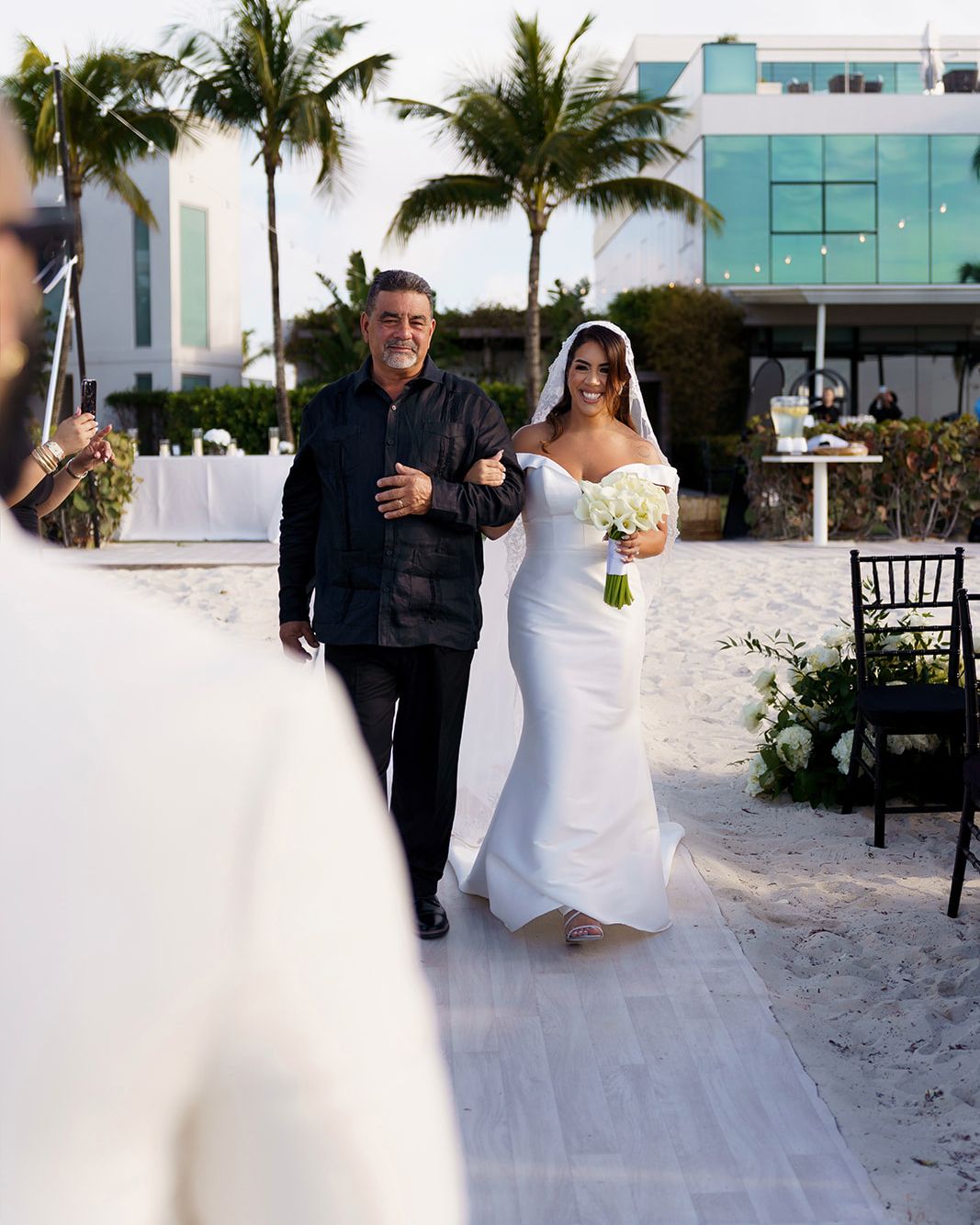 Bride in white off-shoulder gown walks down beach aisle with escort at outdoor wedding ceremony near palm trees.