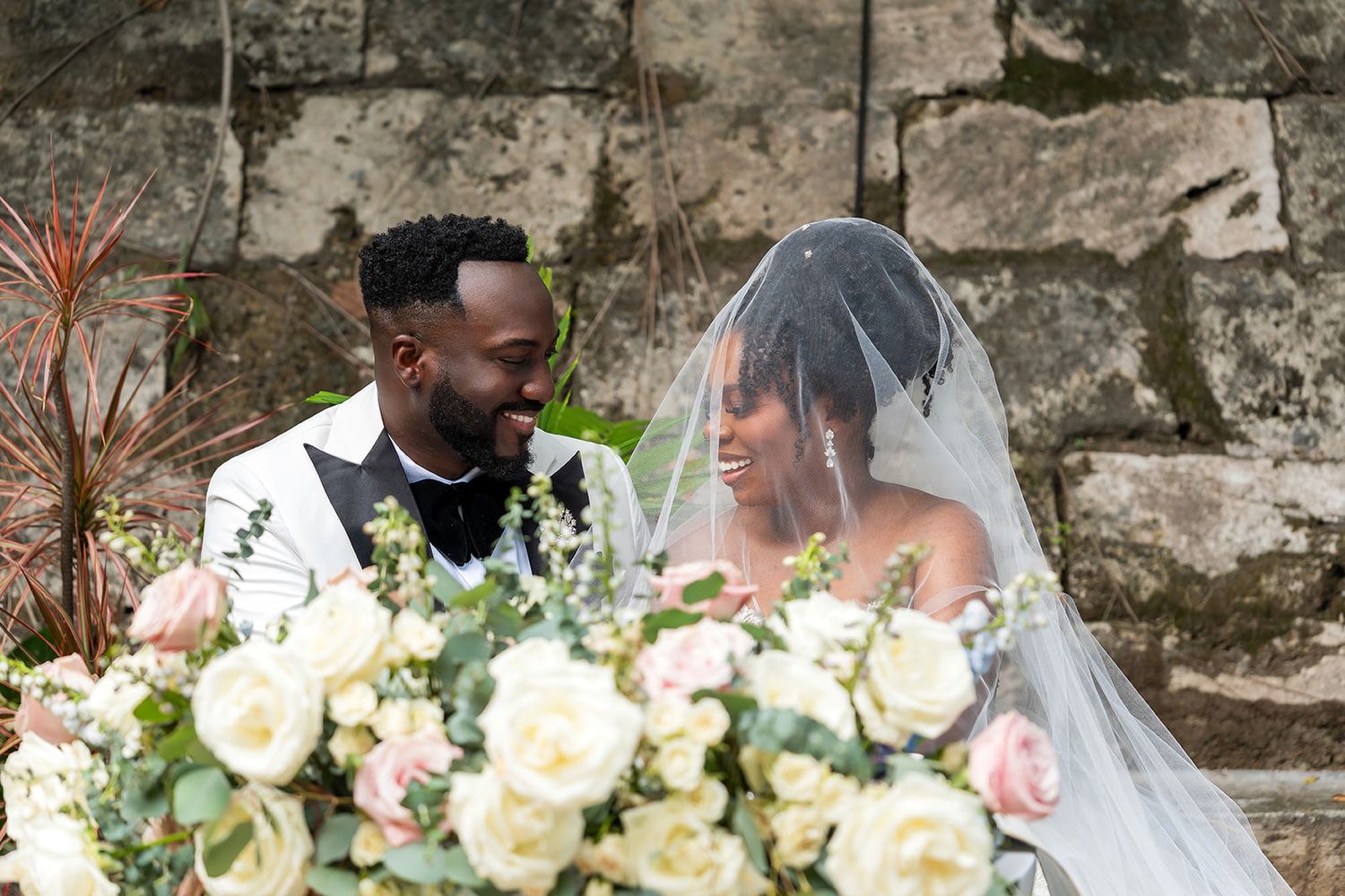 A bride and groom share an intimate moment surrounded by white roses, the bride wearing a veil at an outdoor stone wedding venue.