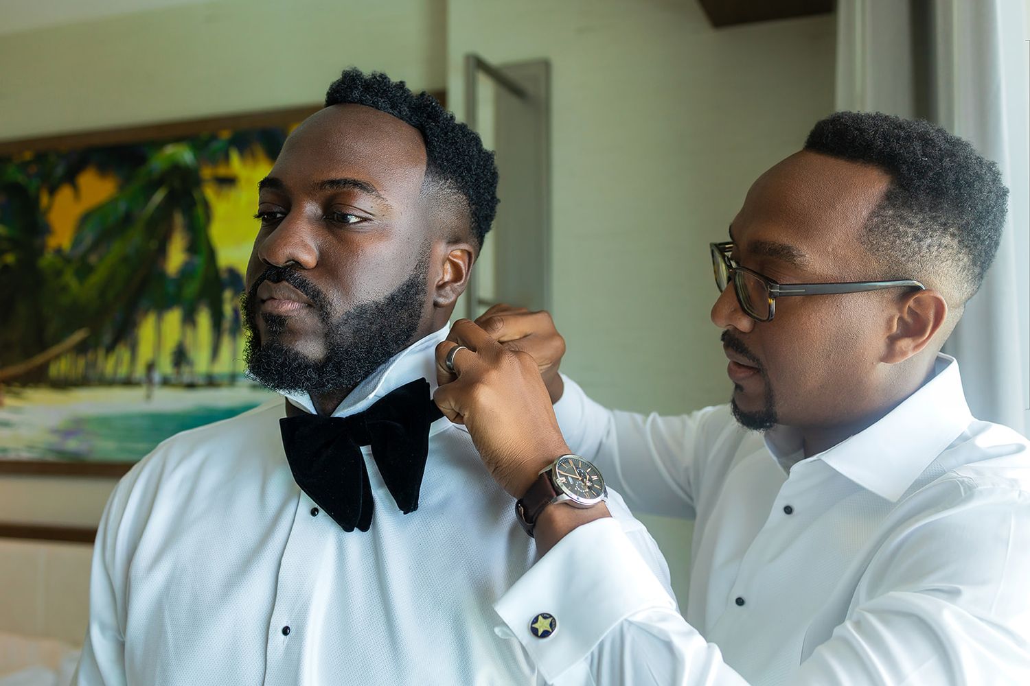 Groom in white dress shirt having black bow tie adjusted by groomsman before wedding ceremony.