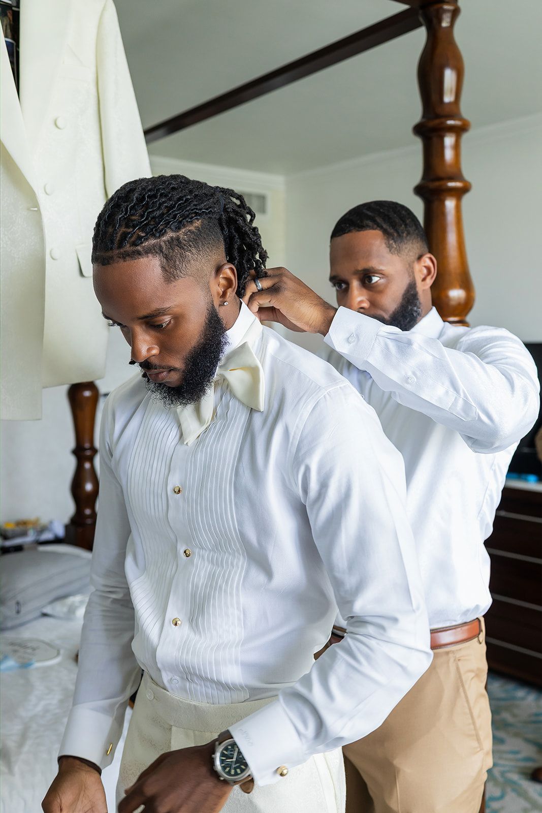 Two well-dressed Black men in white dress shirts preparing for a wedding, one helping adjust the other's collar.