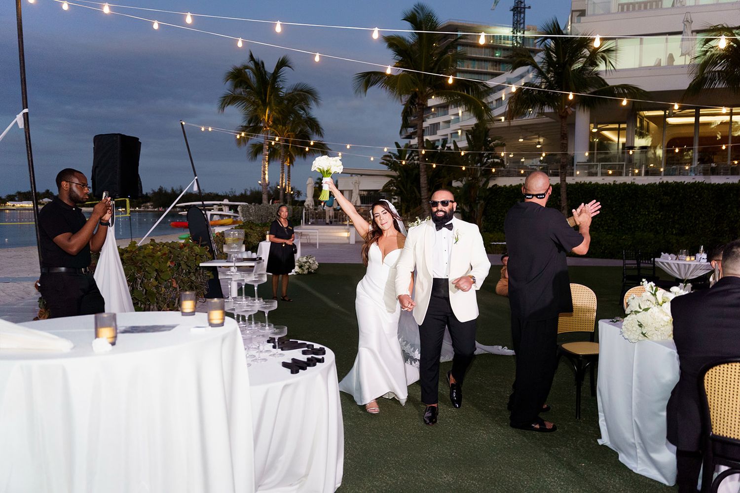 Bride and groom make grand entrance at outdoor waterfront wedding reception with string lights and palm trees.