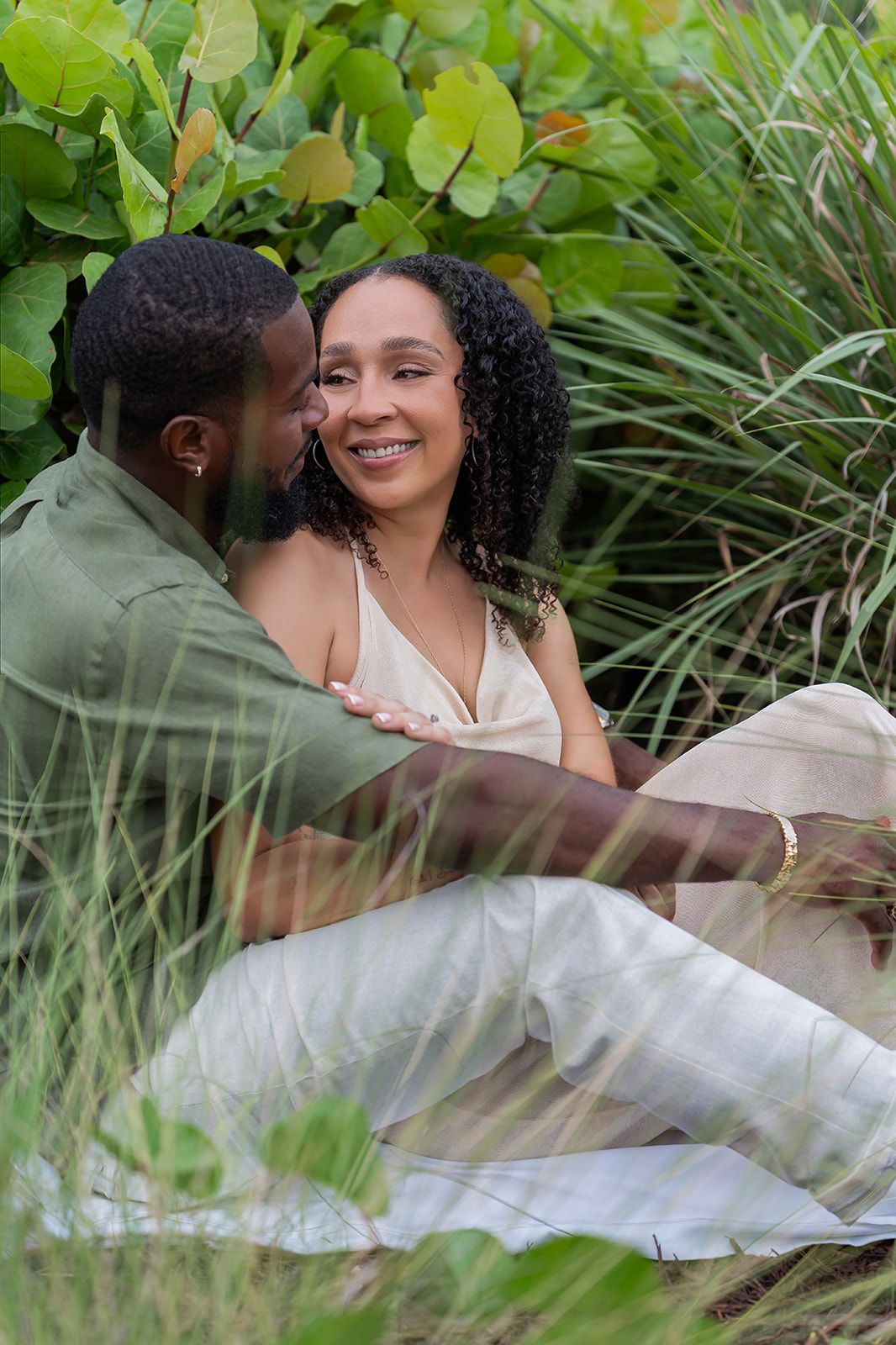 A smiling couple sits together among lush green tropical plants, sharing an intimate moment outdoors.