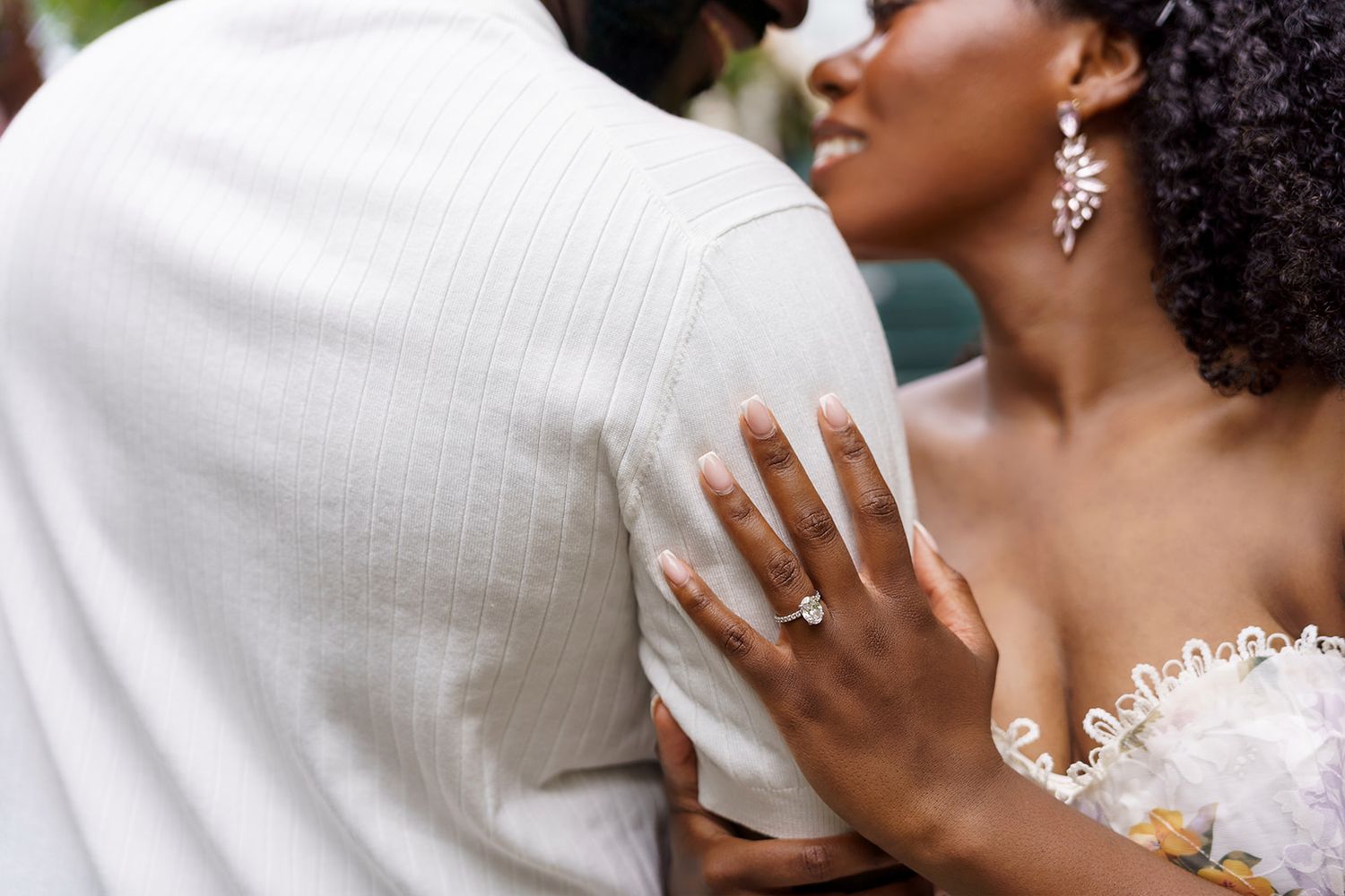 Bride wearing diamond engagement ring and earrings embracing groom in white suit at wedding ceremony.