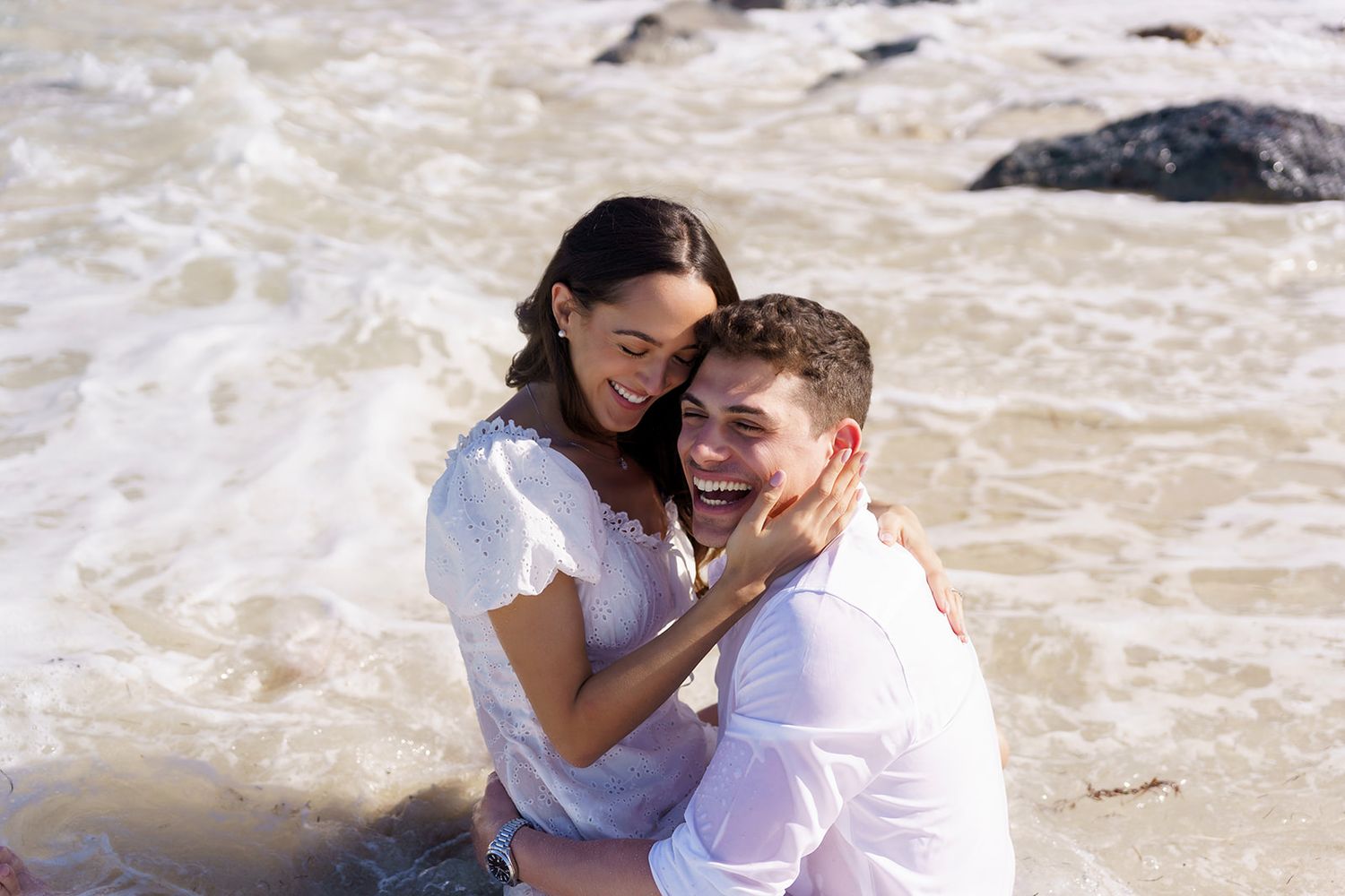 Happy couple embracing and laughing together while sitting in ocean waves on a sunny beach day.