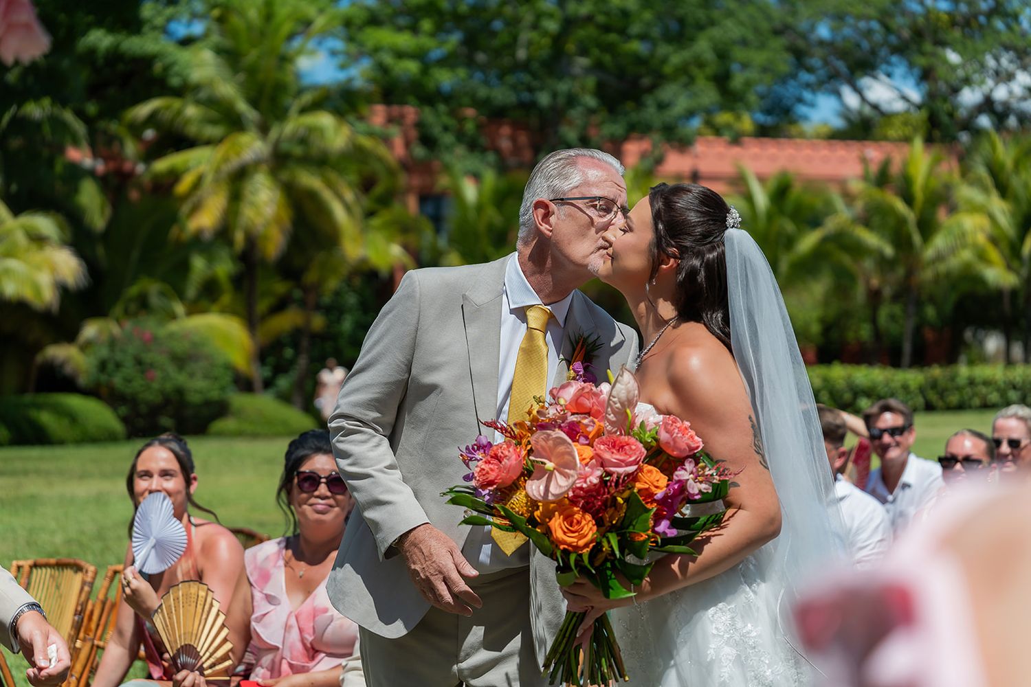 Bride in white gown kisses older man in tan suit at outdoor wedding ceremony with tropical greenery.