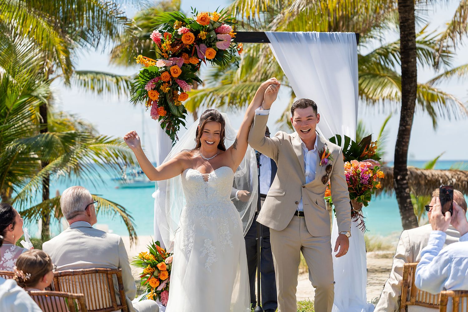 Joyful newlyweds celebrate at outdoor beach wedding ceremony with tropical floral arch and palm trees.