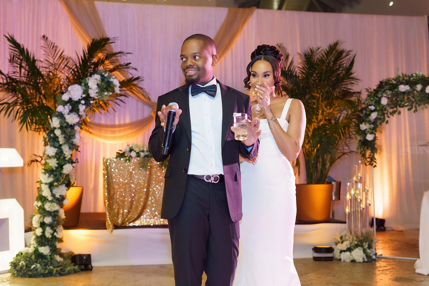 Bride and groom share a toast during wedding reception, surrounded by floral arrangements and warm golden lighting.