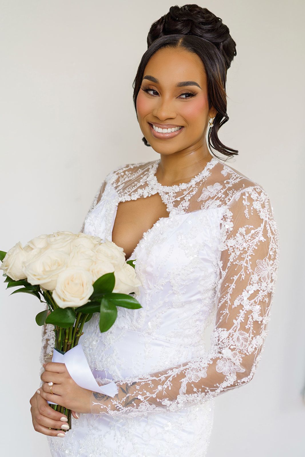 Smiling bride in lace wedding gown holds white rose bouquet with elegant updo hairstyle.