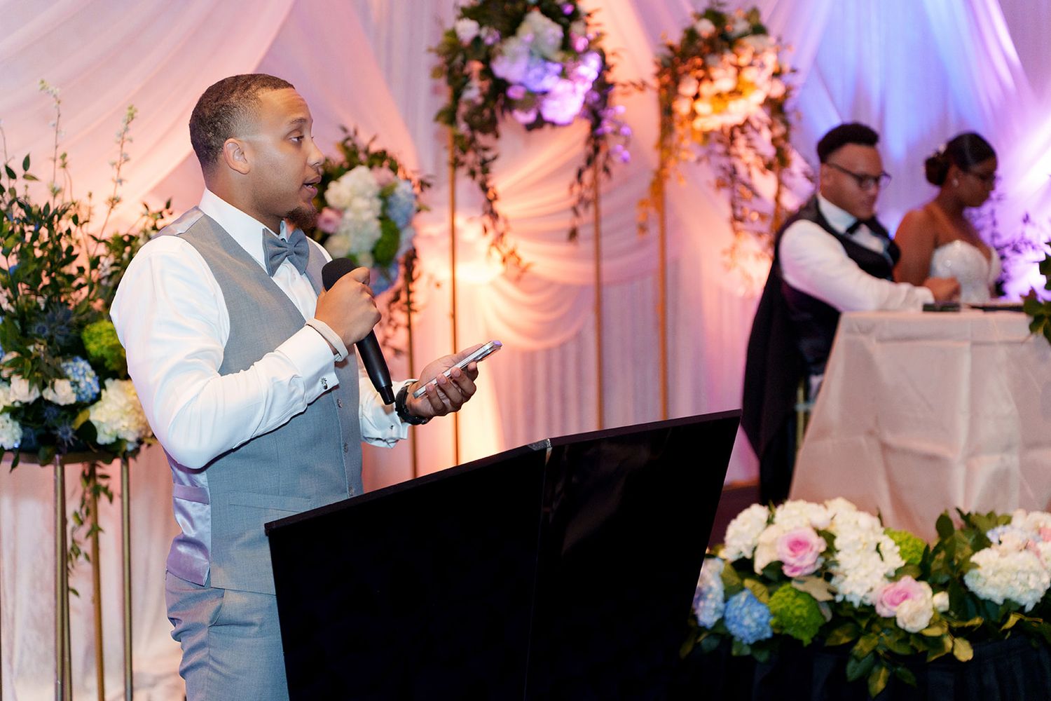 Man in gray vest speaks at wedding reception podium with floral arrangements and couple visible in background.