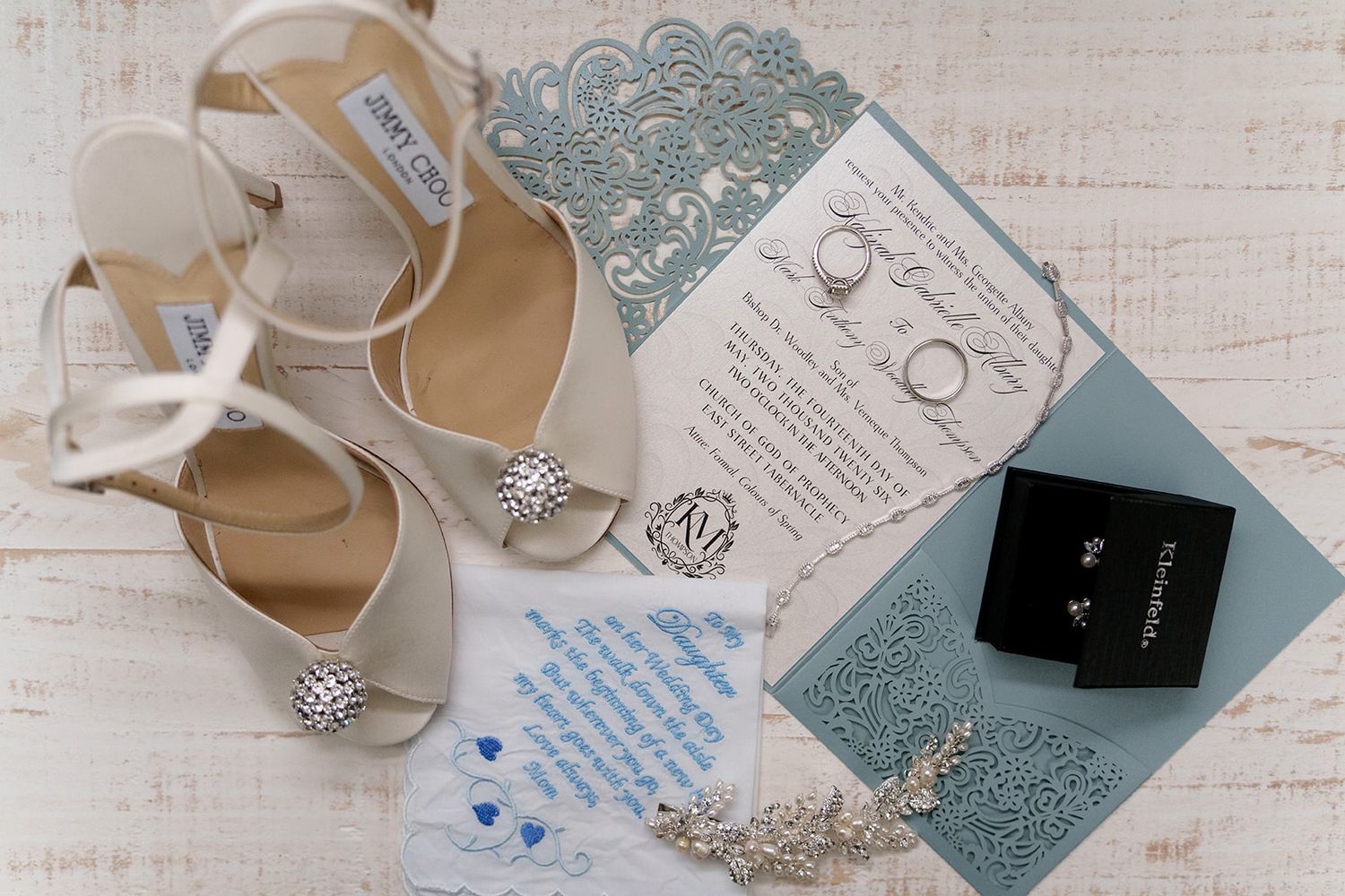 Bridal flat lay featuring Jimmy Choo heels, blue laser-cut invitation, pearl earrings, and delicate jewelry on white wood.