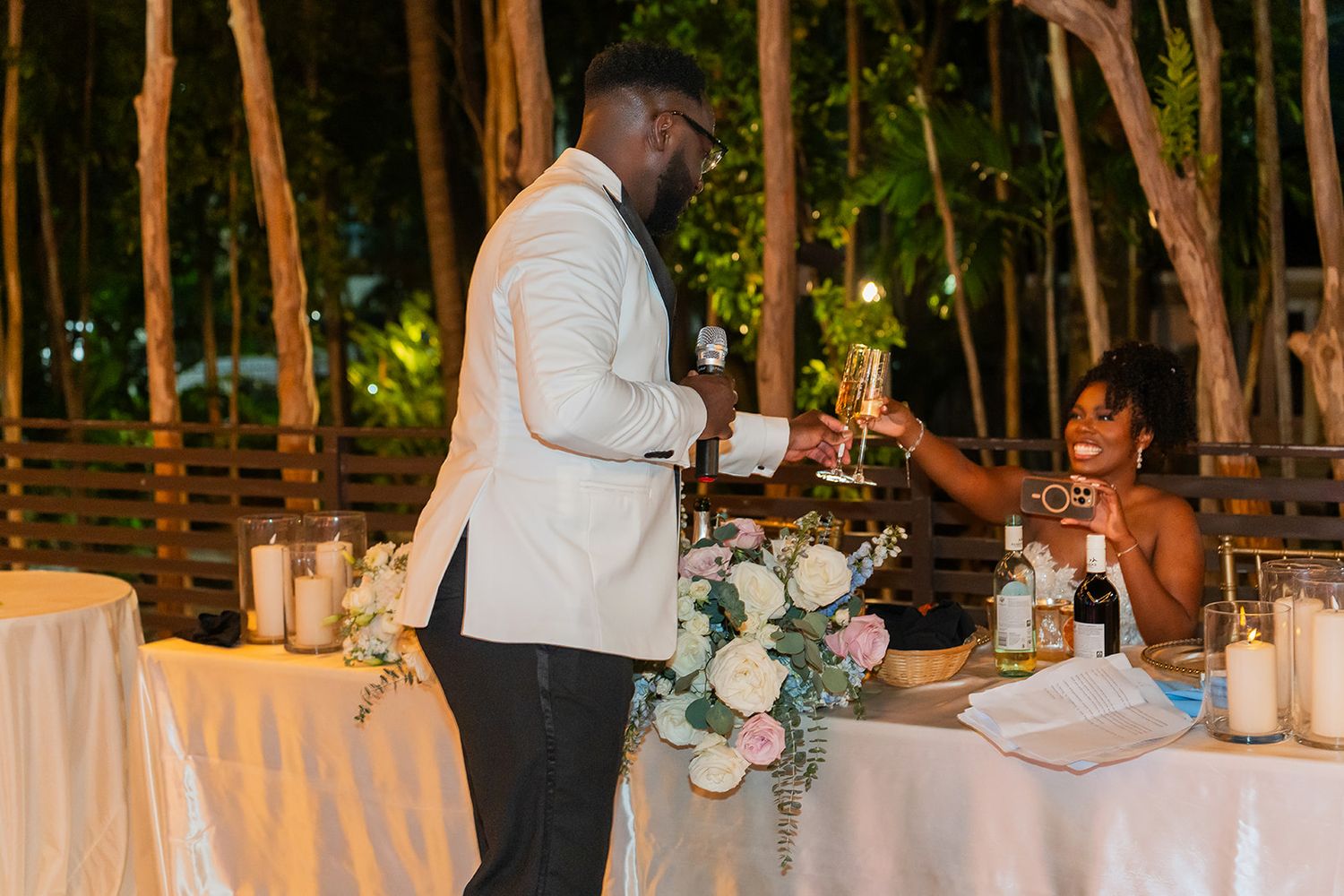 Groom in white tuxedo toasting with bride at elegant outdoor wedding reception with floral centerpiece.