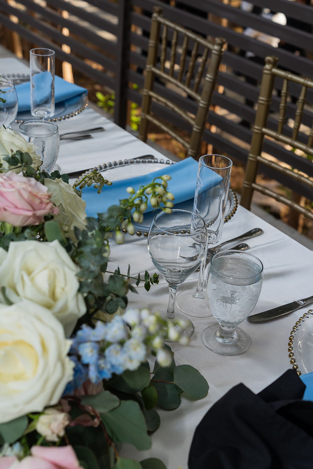Elegant wedding reception table set with blue napkins, crystal glassware, and fresh floral centerpieces.