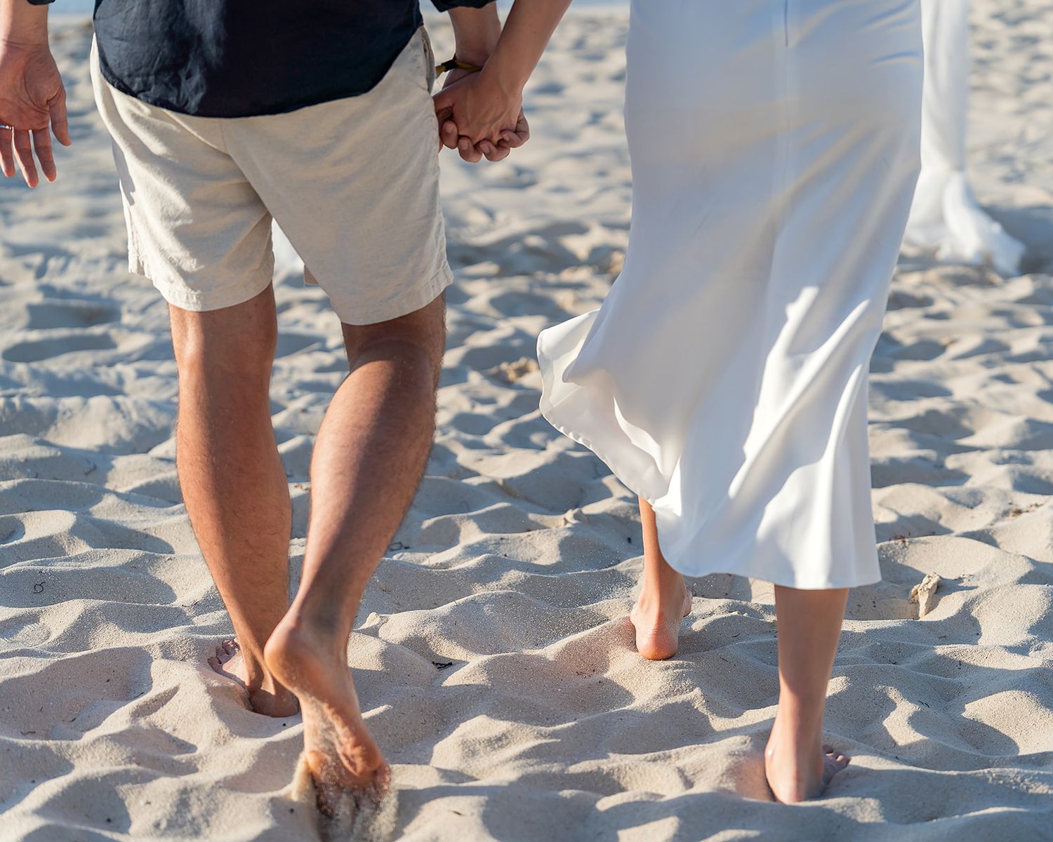 Barefoot newlywed couple holding hands walking on sandy beach in wedding attire.