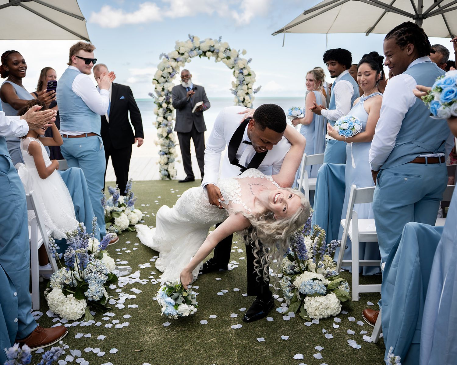 Groom dips bride in romantic kiss at outdoor wedding ceremony with blue-themed wedding party and floral arch.