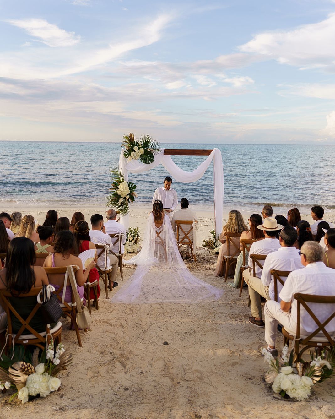 A romantic beach wedding ceremony with guests seated on wooden chairs facing a couple at a white floral arch by the ocean.