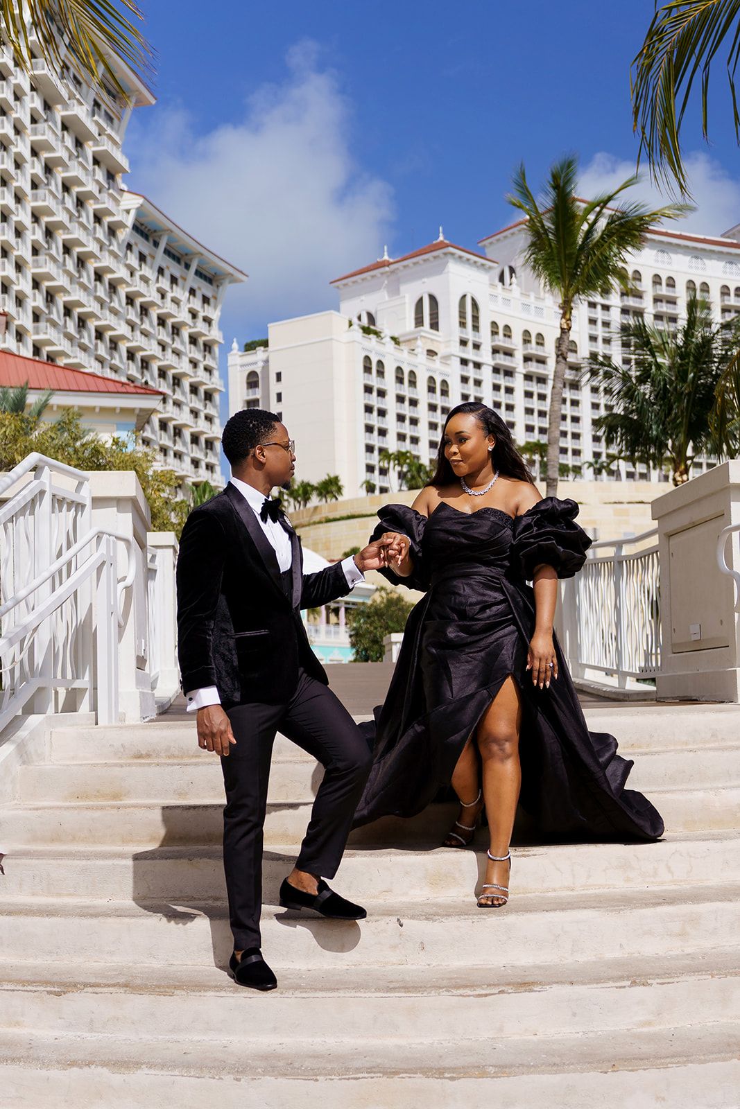 Elegant couple in black formal attire posing on white stairs at a tropical resort with palm trees.