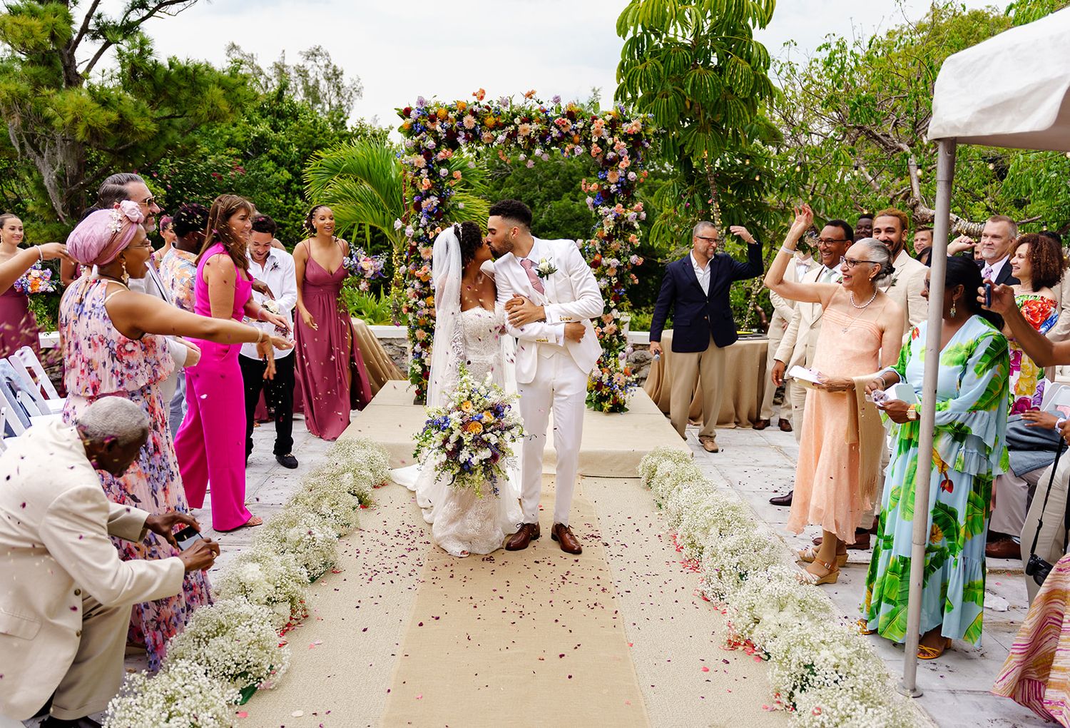 Couple shares first kiss at outdoor wedding ceremony while guests celebrate with flower petals along floral-lined aisle.
