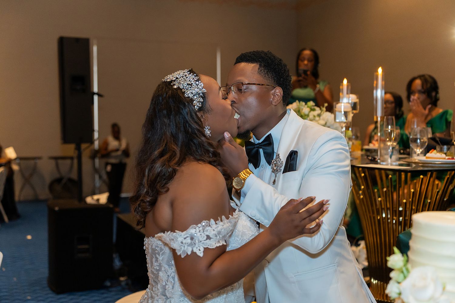 Bride and groom share a kiss during wedding reception cake cutting ceremony, bride wearing lace gown and floral crown.