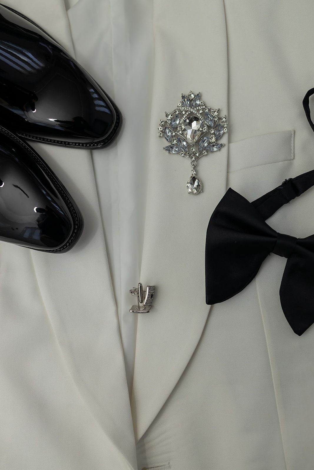White tuxedo jacket with crystal brooch, black bow tie, cufflink, and patent leather dress shoes for formal wear.
