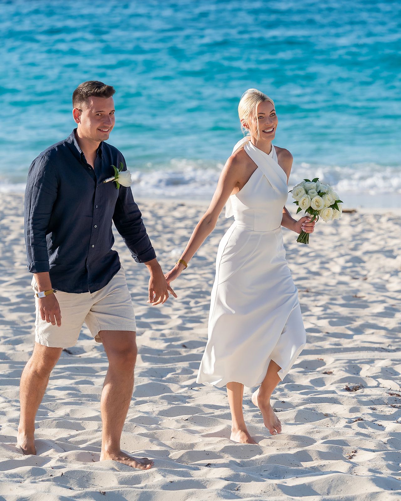 Happy couple walking barefoot on a sandy beach, bride in white dress holding bouquet, groom in navy shirt.