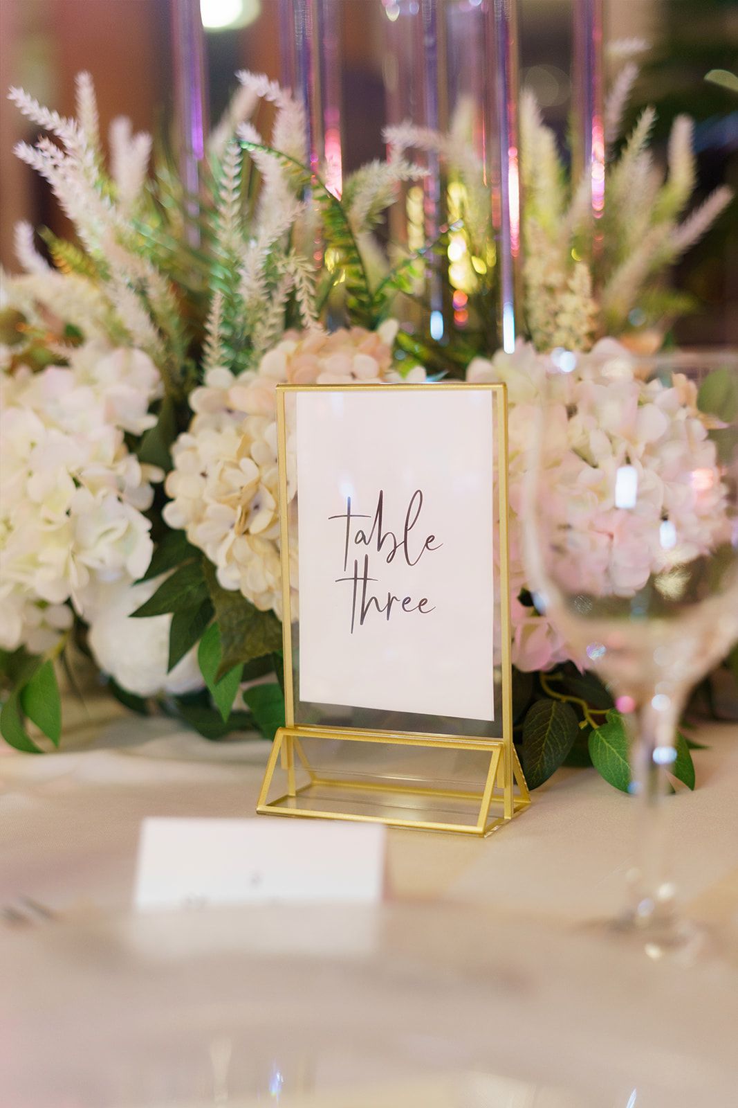 Gold-framed table three sign at elegant wedding reception with white hydrangea floral arrangements.