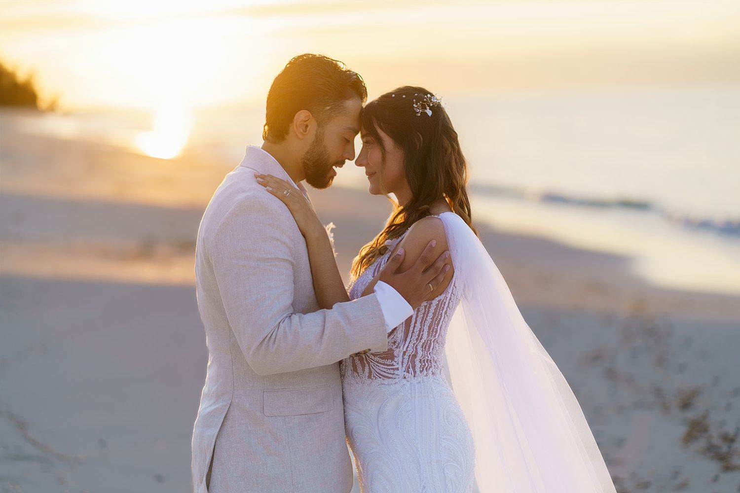 Bride and groom share a romantic sunset embrace on a beach, with golden light glowing behind them.