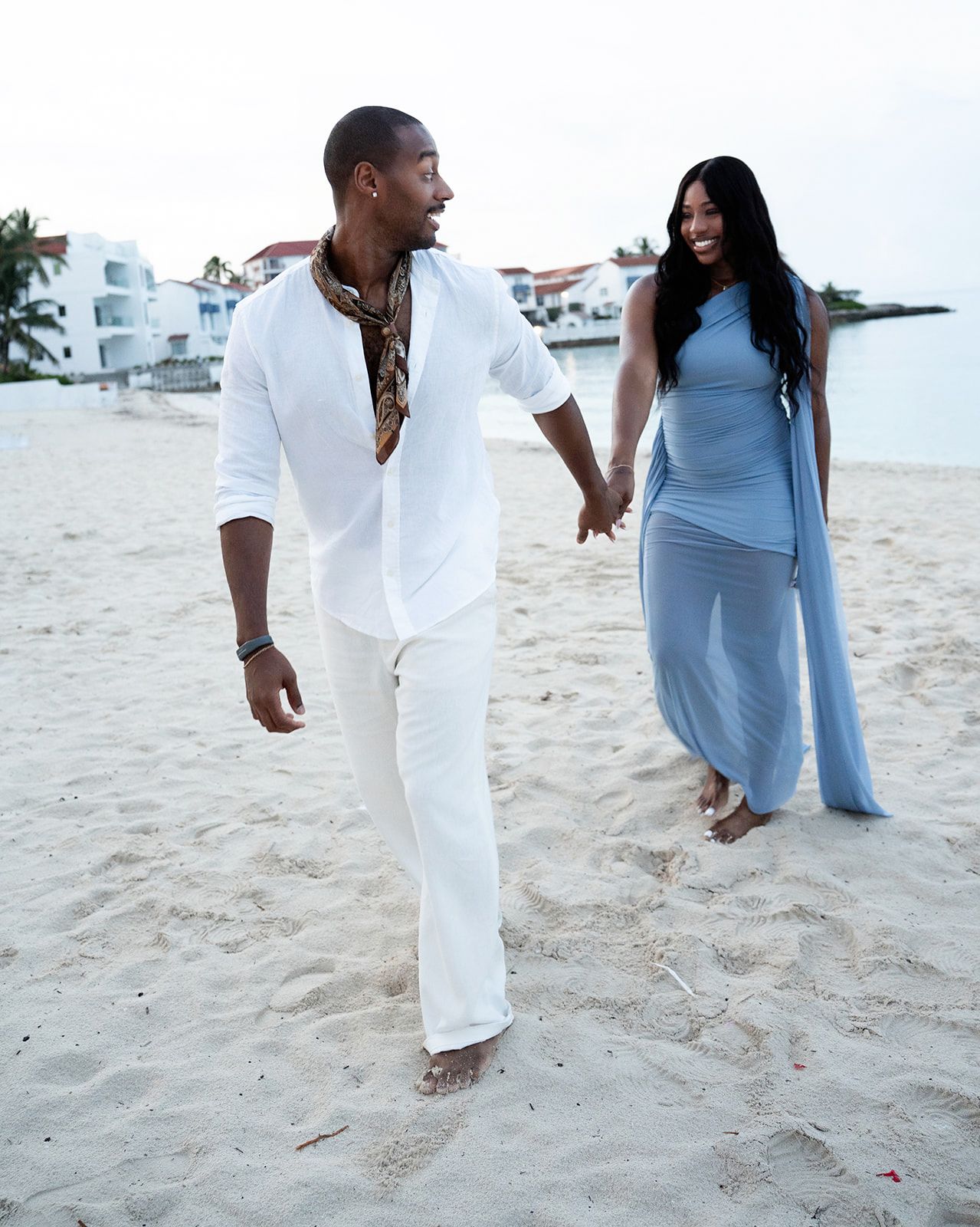 Couple holding hands walking barefoot on a sandy beach, man in white linen outfit, woman in flowing blue dress.