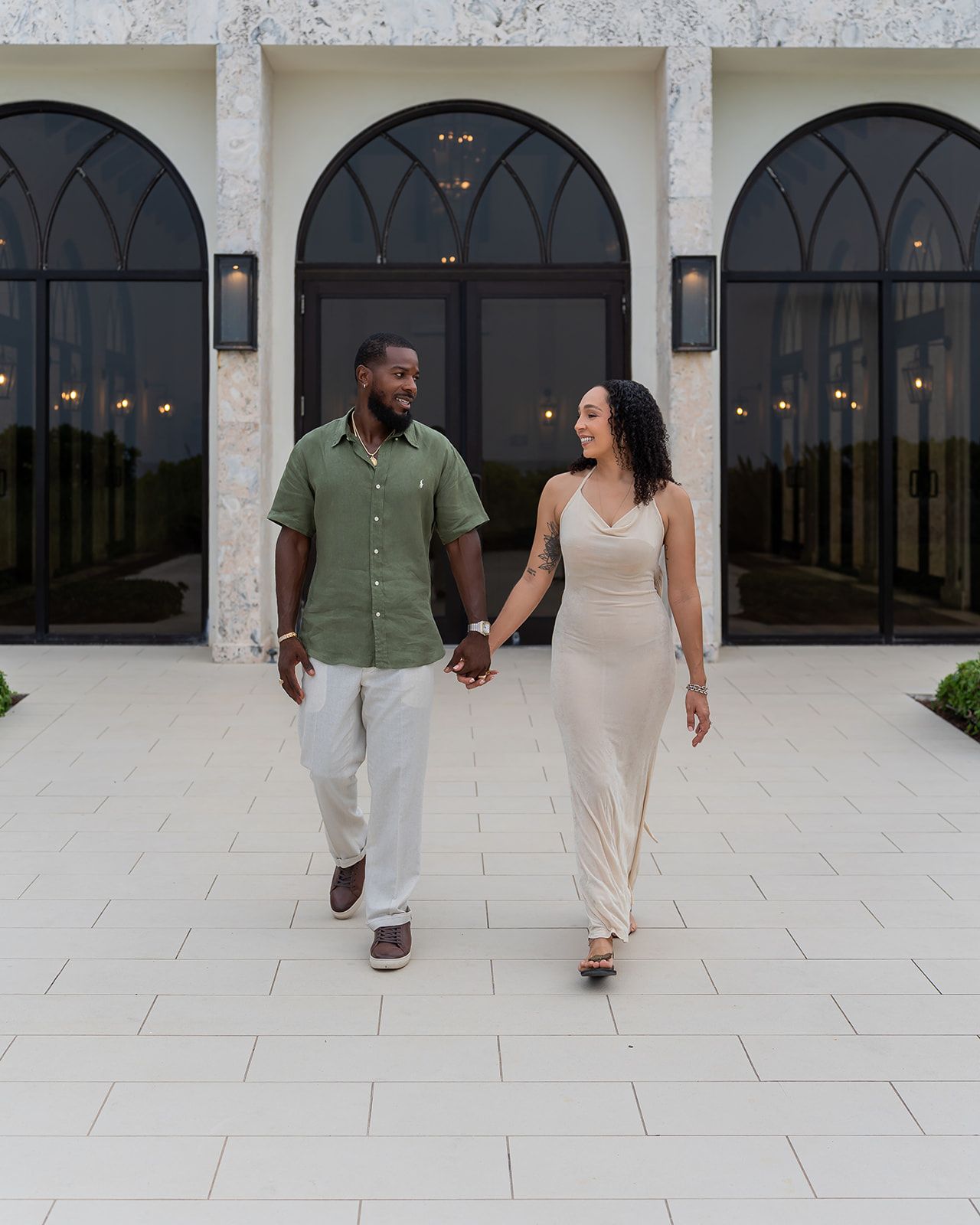 Couple holding hands walking in front of elegant arched building with black windows at dusk.