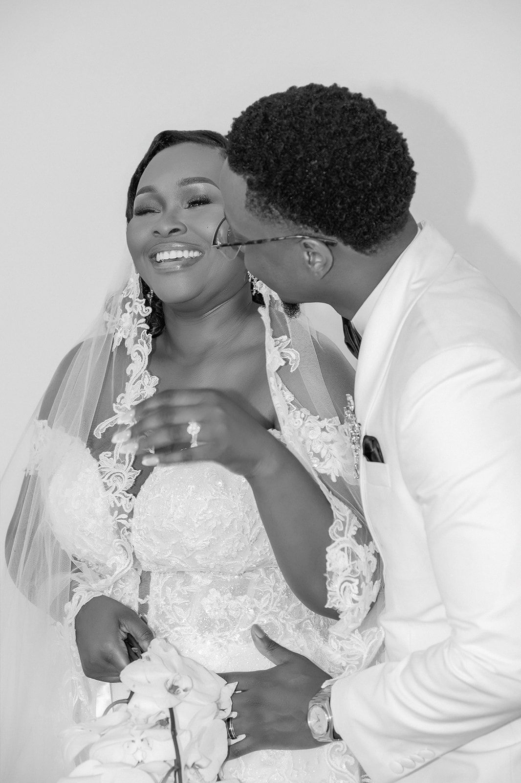 Black and white photo of a groom kissing his bride on the cheek as she smiles in a lace wedding dress and veil.