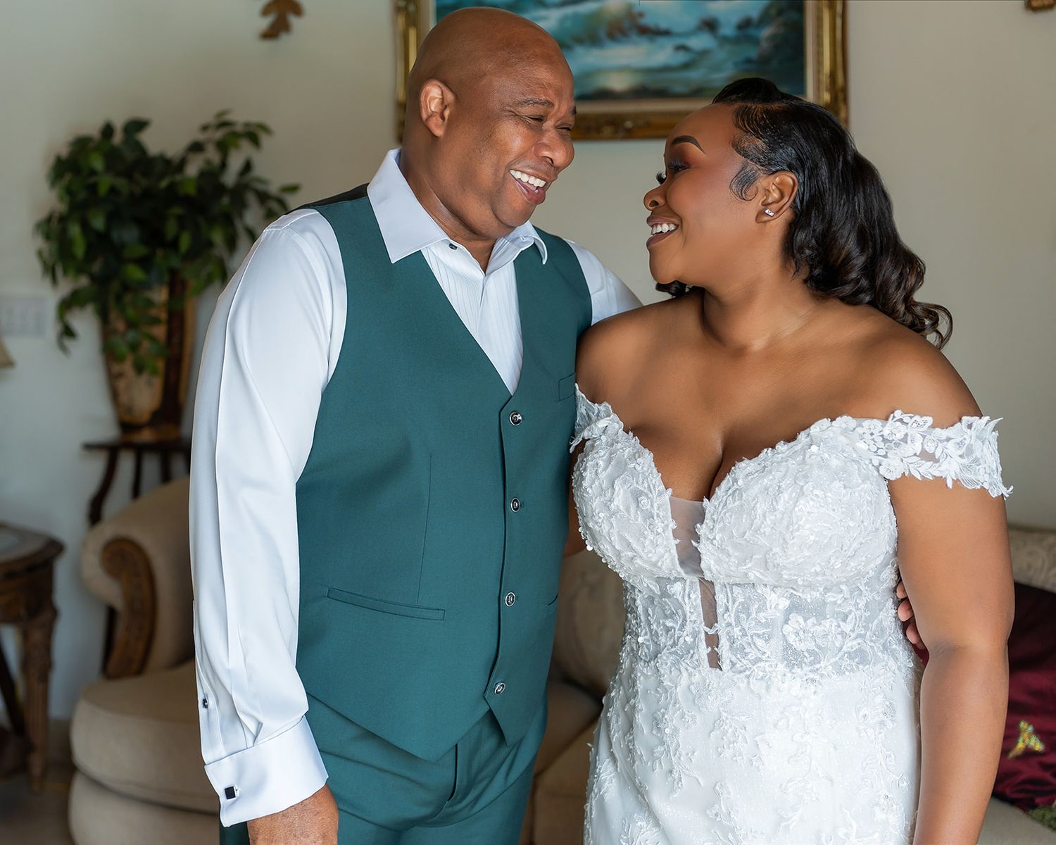 A joyful couple shares a loving glance, bride in lace off-shoulder gown and groom in teal vest, during wedding day preparations.