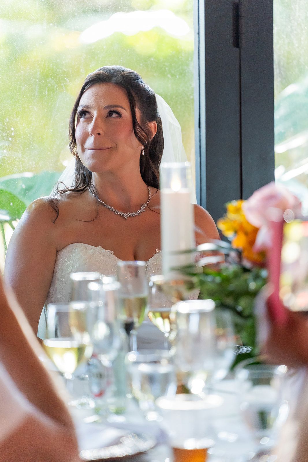 Bride in white strapless gown and veil sits at wedding reception table with candles and champagne glasses.