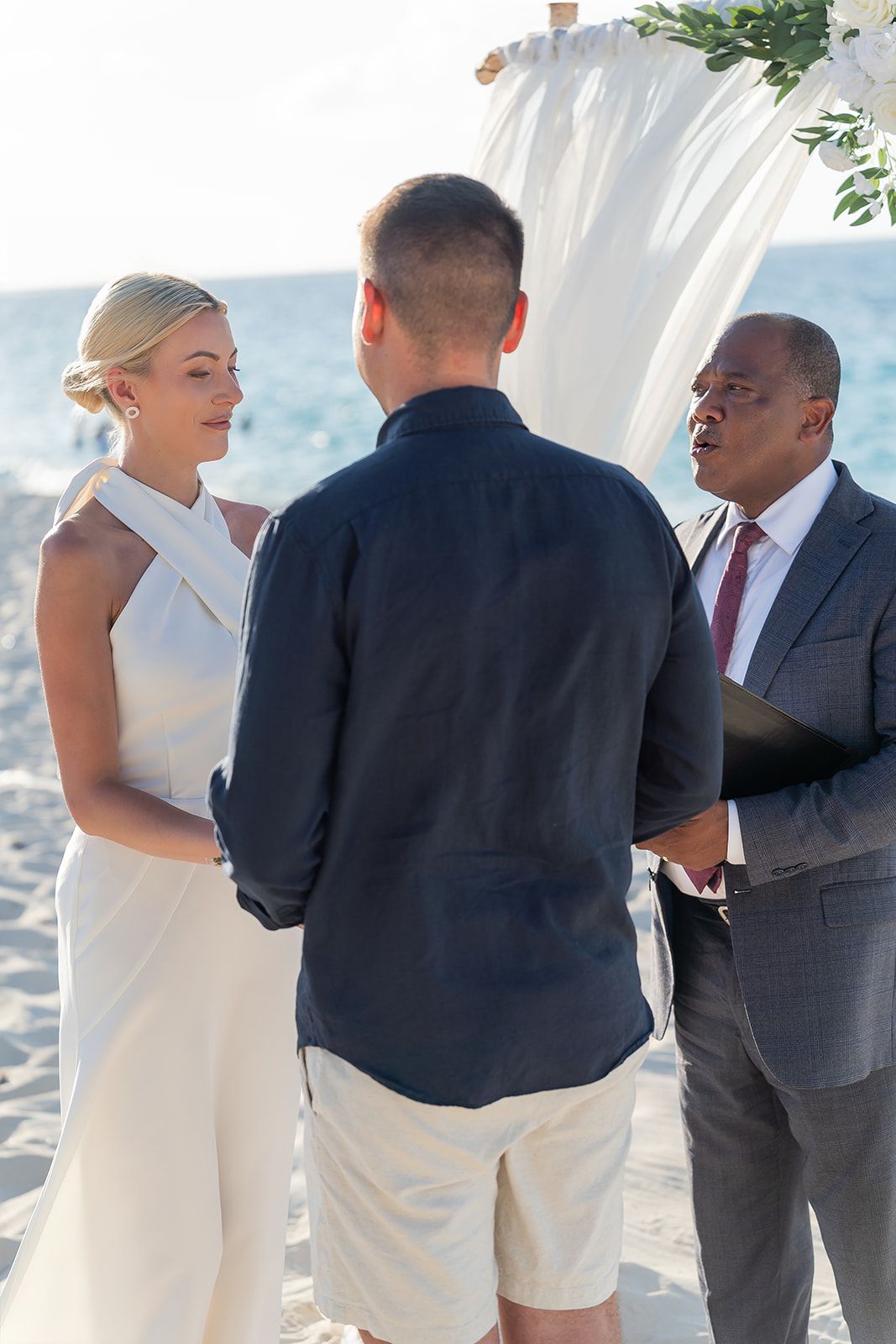 Couple exchanging vows on a beach wedding ceremony with an officiant under a white draped arch.