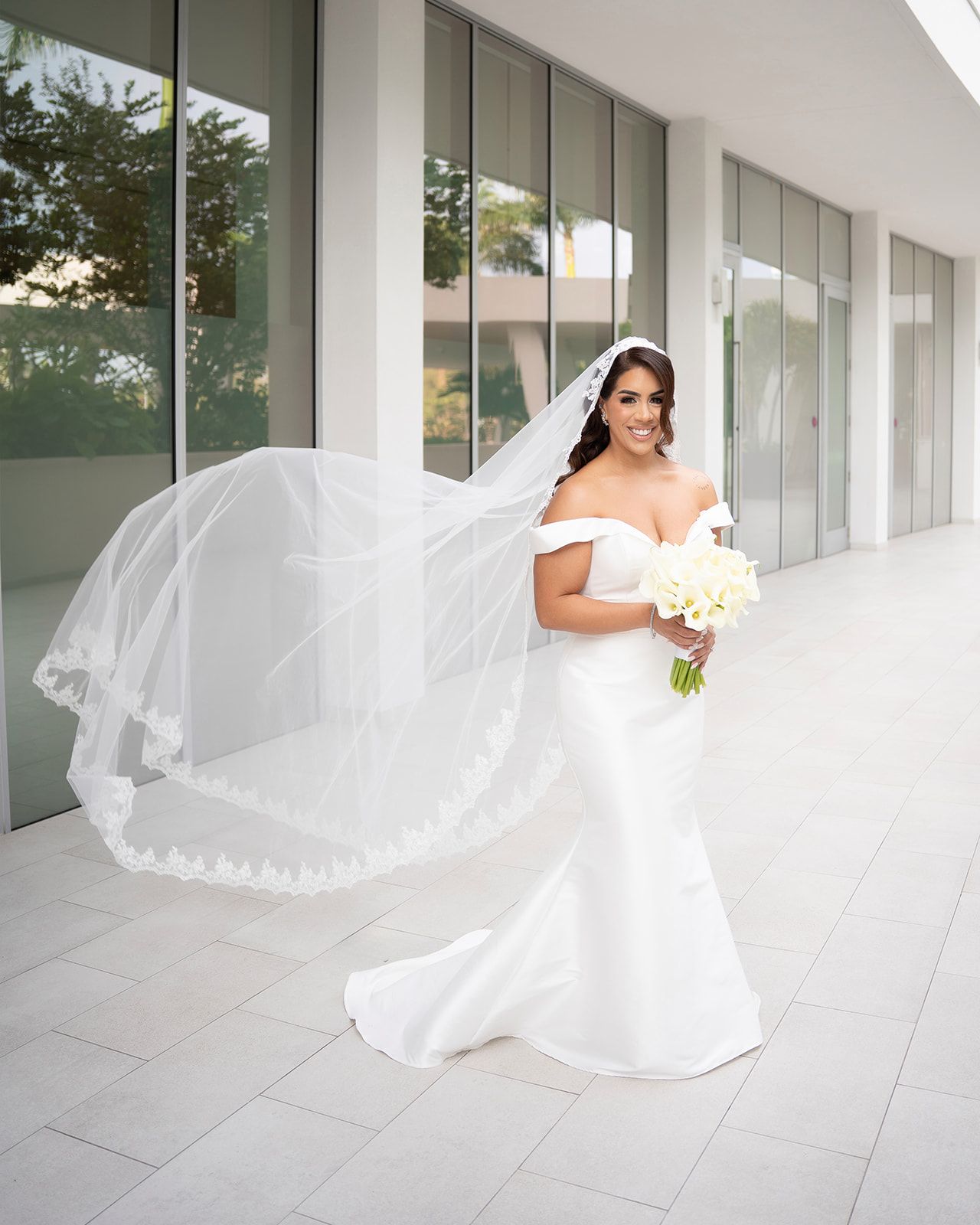 Bride in off-shoulder mermaid gown with flowing lace veil holding white bouquet, posing outdoors at modern venue.