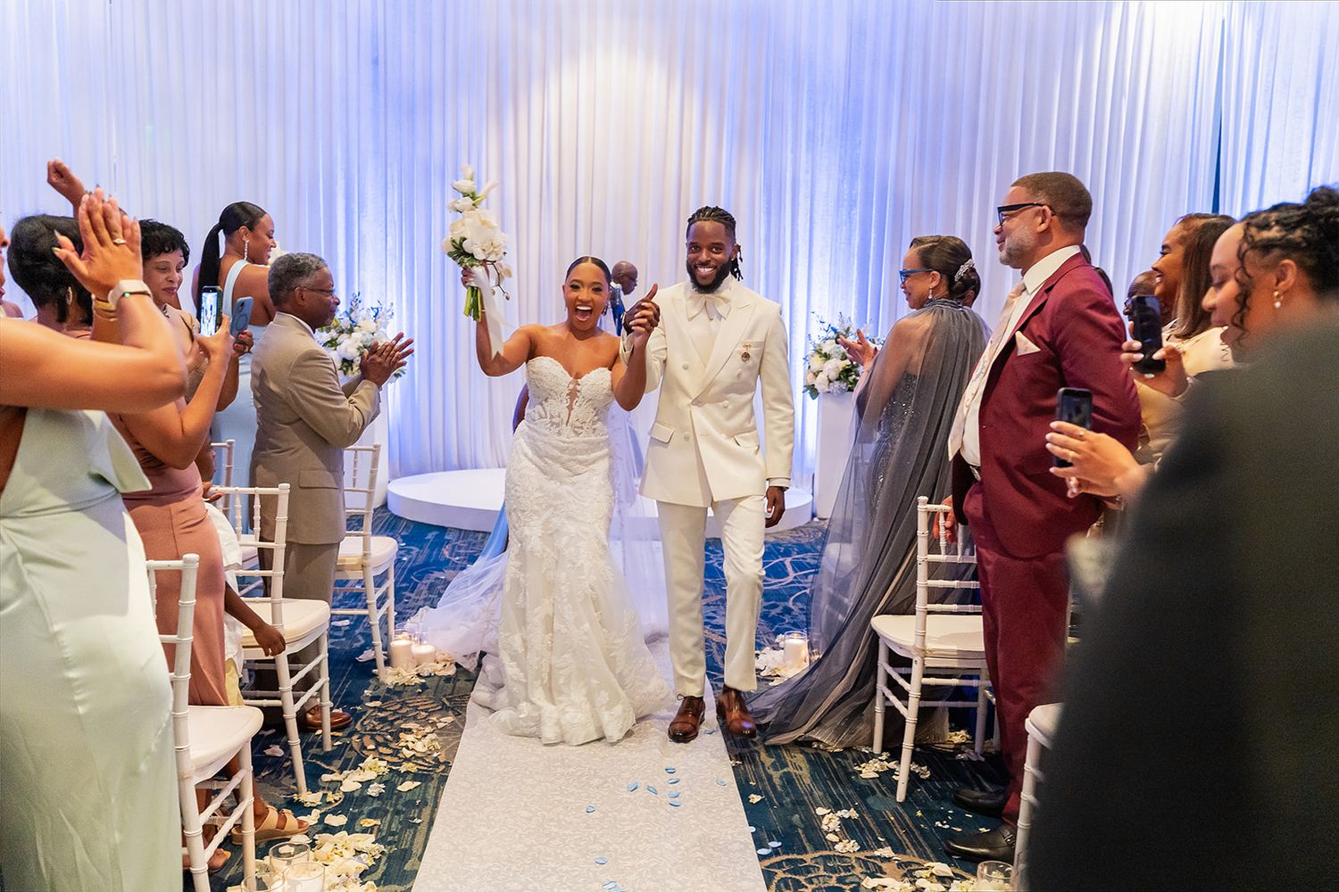 Joyful newlyweds walk down the aisle after wedding ceremony, guests celebrating with confetti in elegant blue venue.