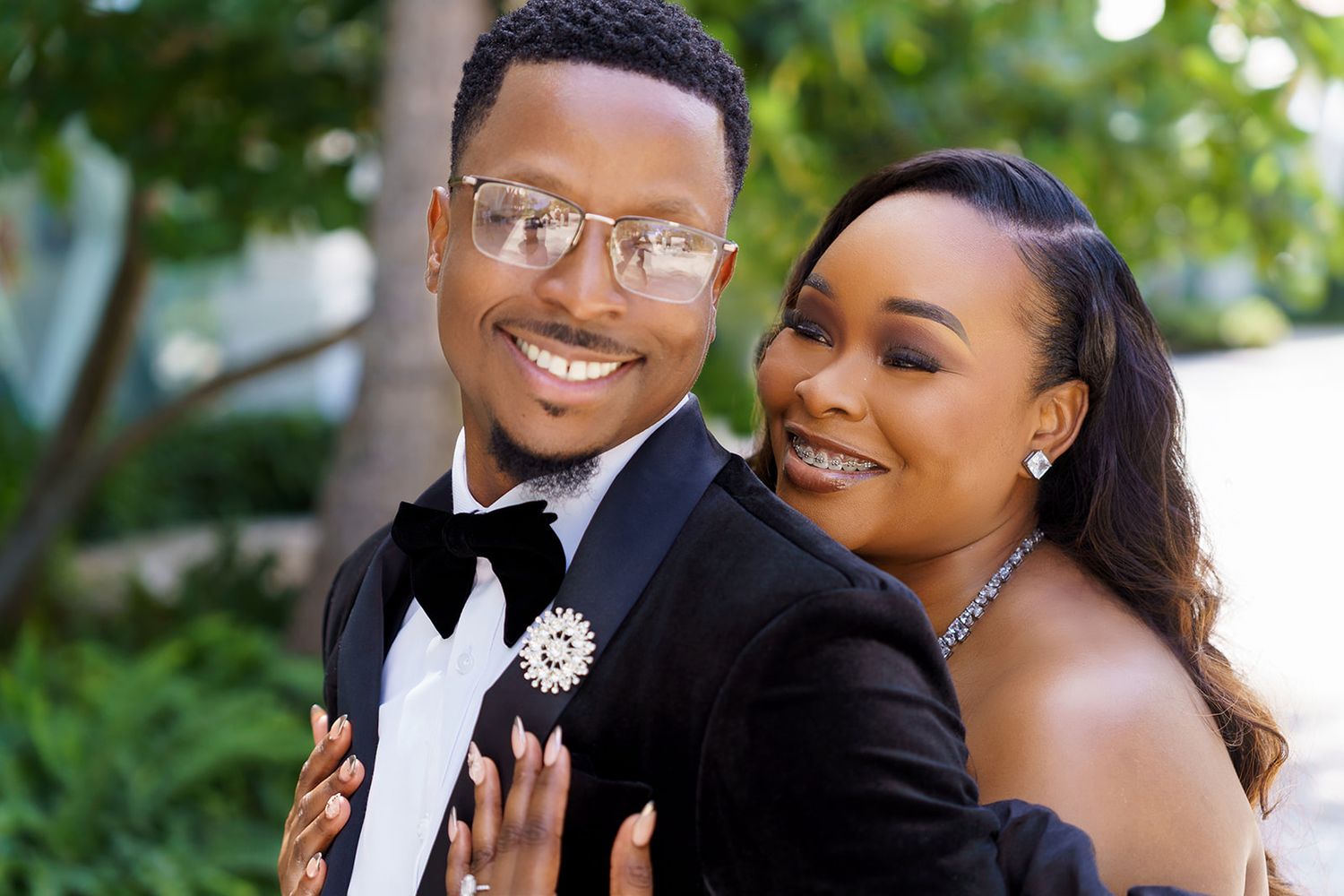 Elegant couple posing together, man in tuxedo with boutonniere, woman in formal attire, smiling outdoors.
