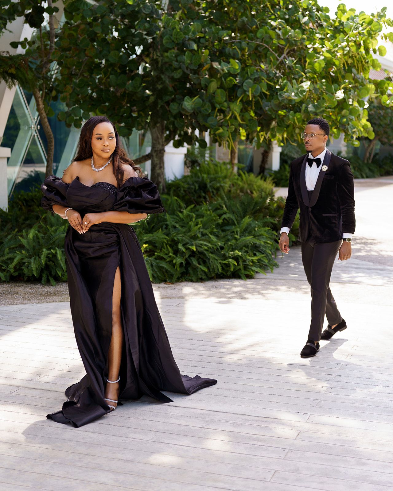 Elegant couple in matching black formal wear pose outdoors, woman in off-shoulder gown with slit, man in velvet tuxedo.