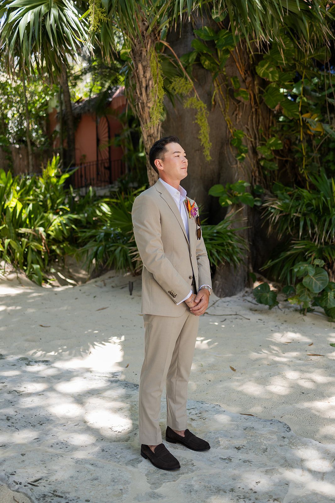 Groom in beige suit with boutonniere stands on sandy beach surrounded by tropical palm trees.