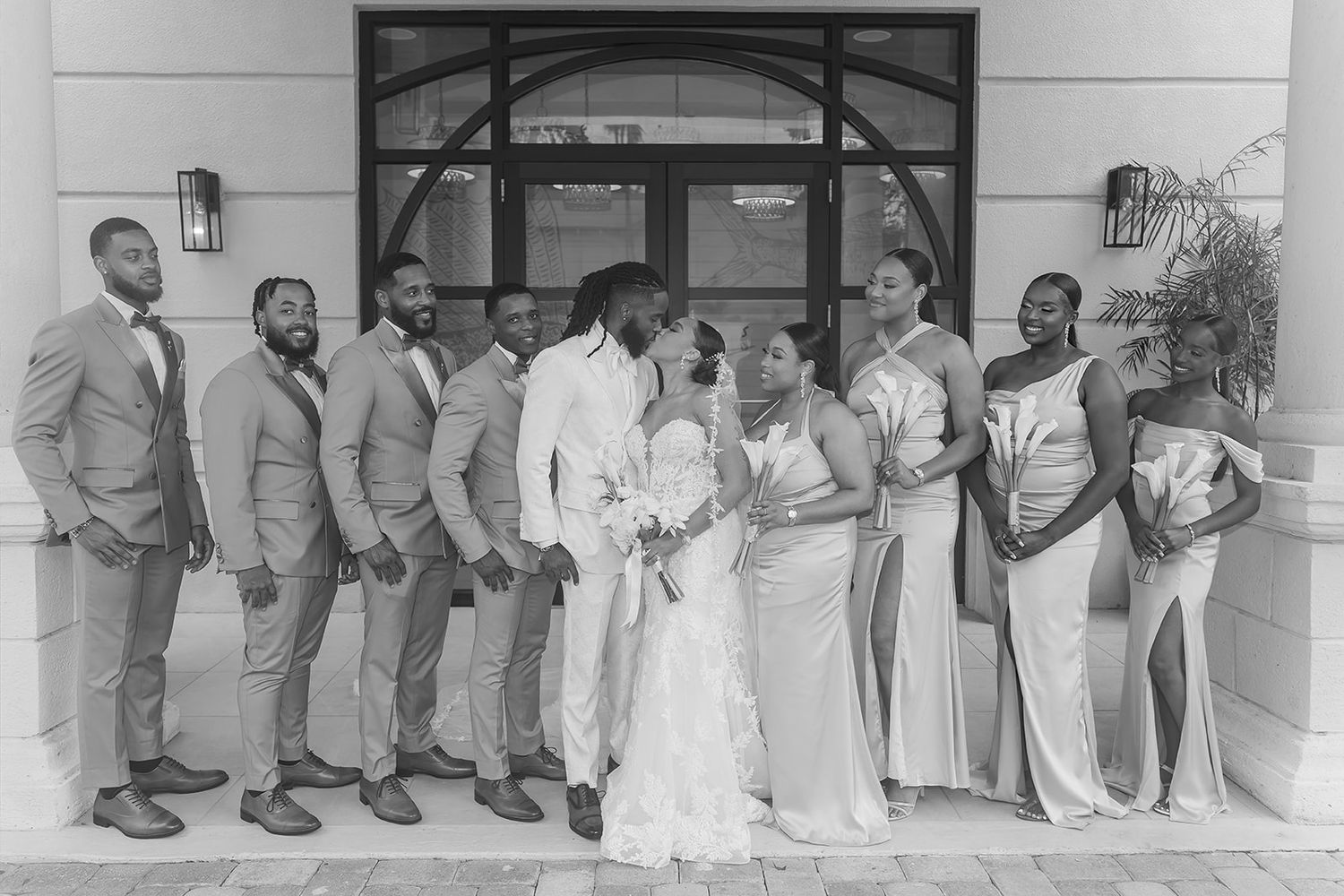 Black and white wedding photo of bride and groom kissing surrounded by bridesmaids and groomsmen outside a building.