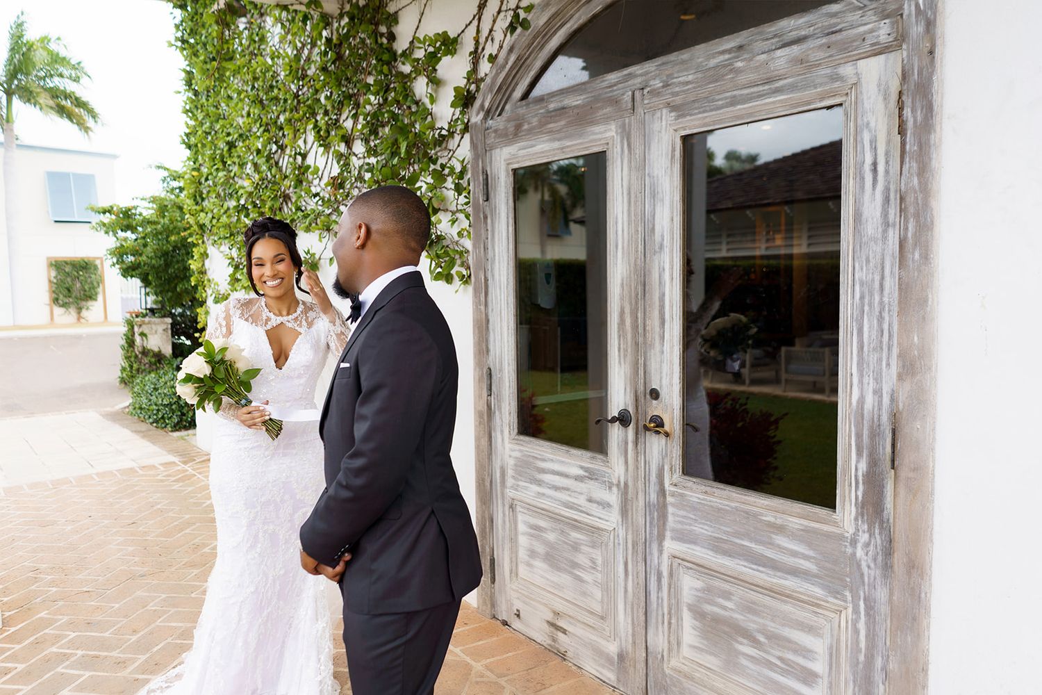 A bride in a lace gown and groom in a tuxedo share a first look moment outside a rustic white door.