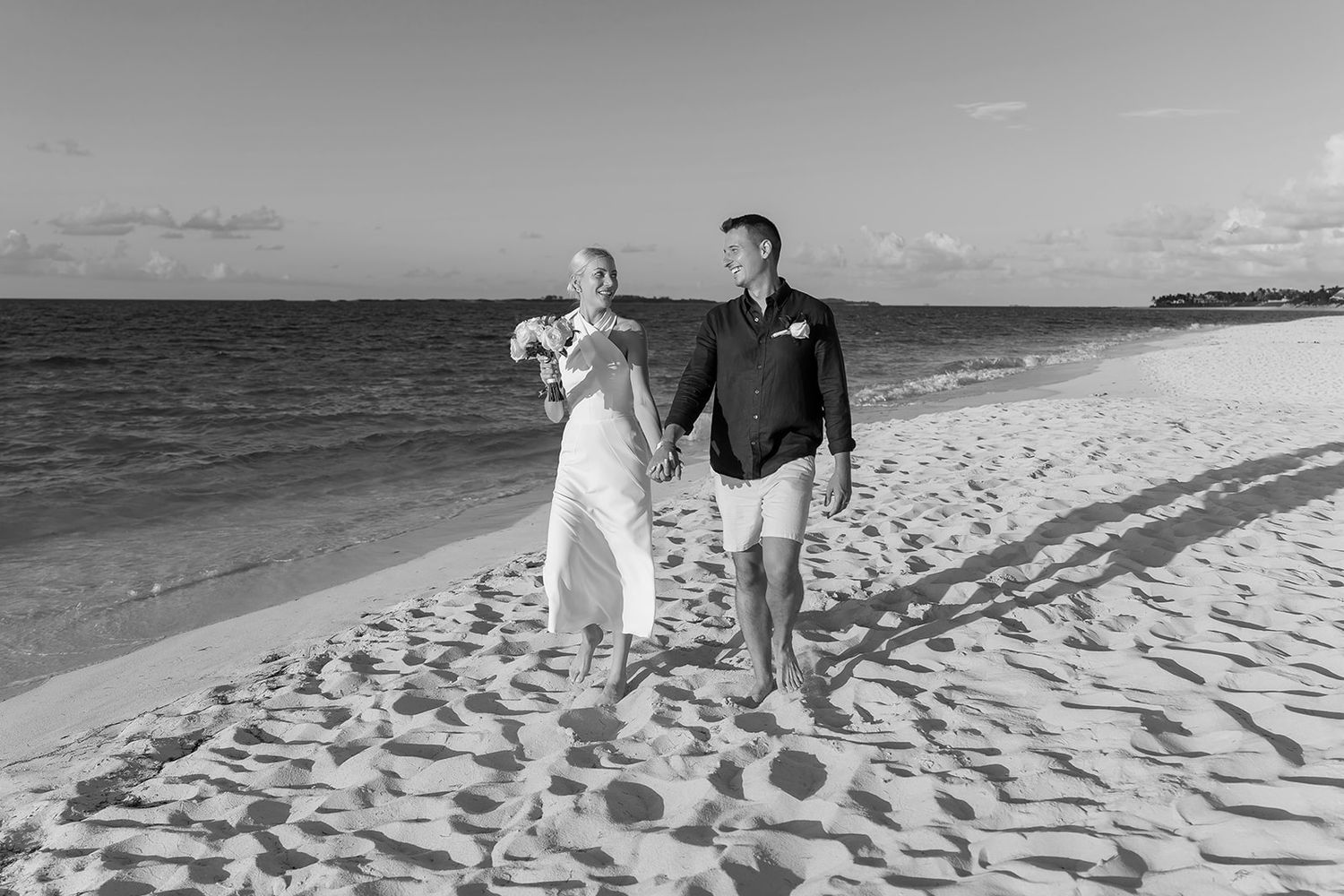 Bride and groom walking hand in hand on a sandy beach in a black and white wedding photo.
