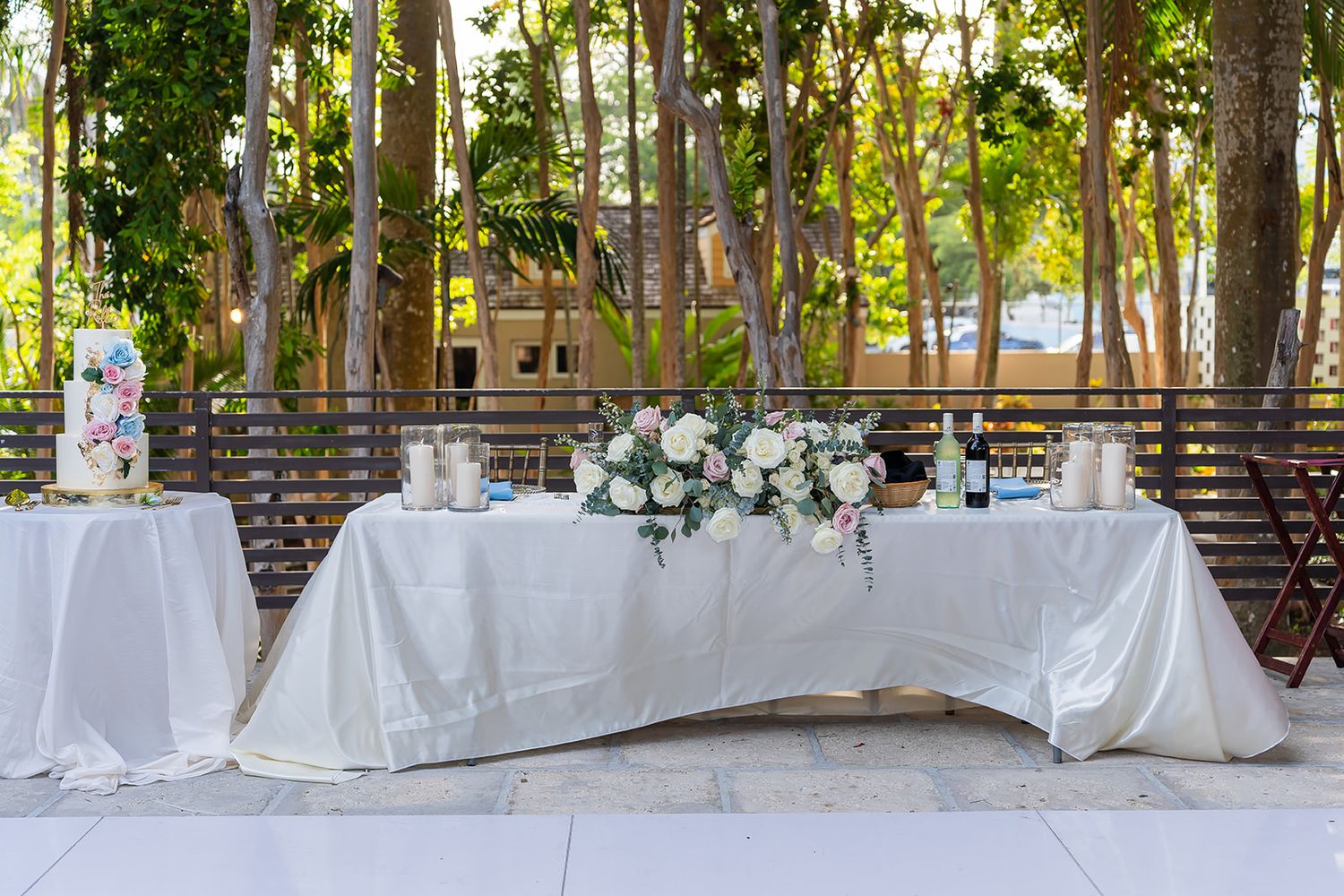 Elegant outdoor wedding reception sweetheart table with white linens, floral centerpiece, and wedding cake.
